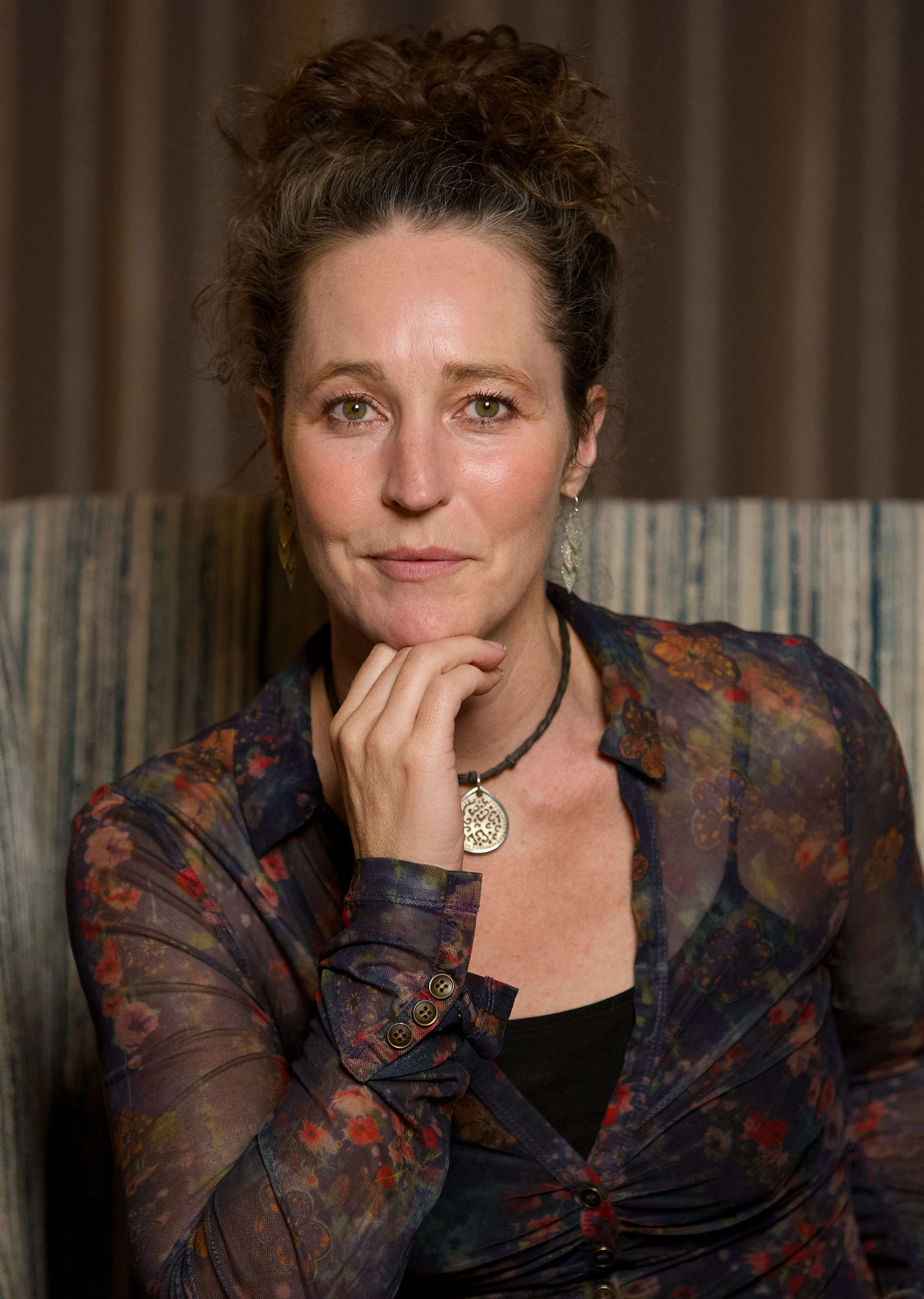 A woman with curly hair and green eyes, wearing a floral-patterned sheer jacket and a black top, sitting in front of a dark brown curtain, resting her chin on her hand, smiling softly at the camera.