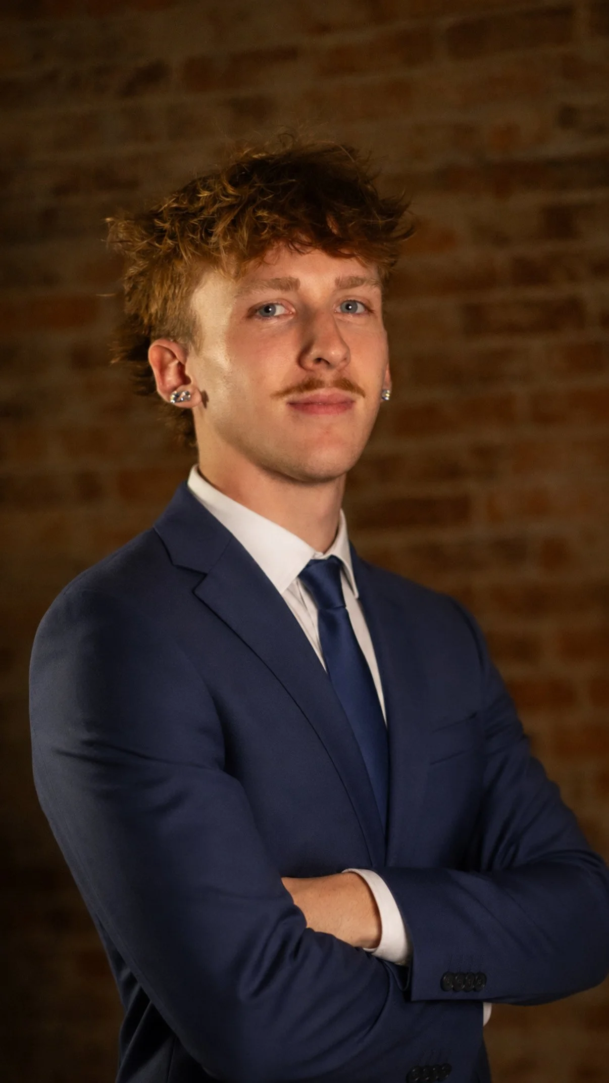 A young man in a navy blue suit with a white shirt and matching navy tie, standing with his arms crossed in front of a brick wall, looking at the camera with a slight smile.