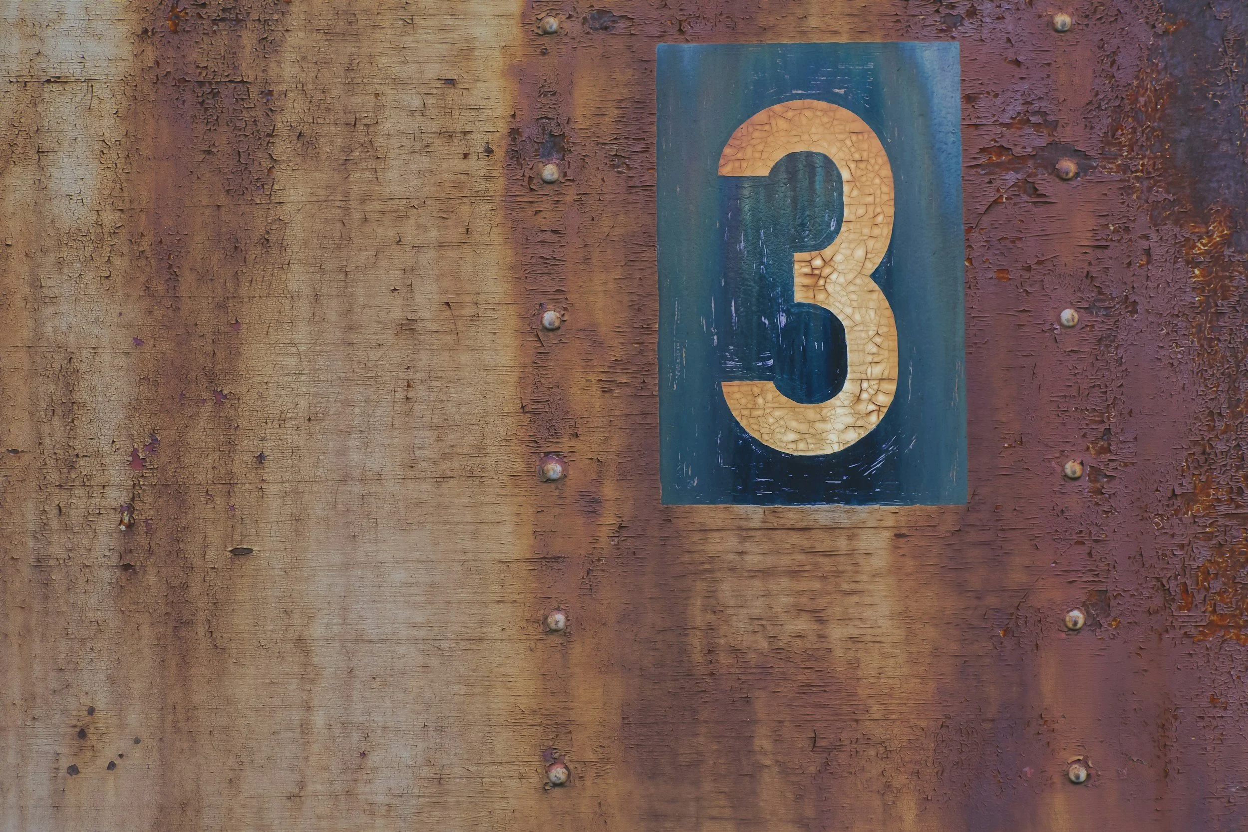 Close-up of a blue metal sign with the number 3, mounted on a weathered, rusted wooden surface.
