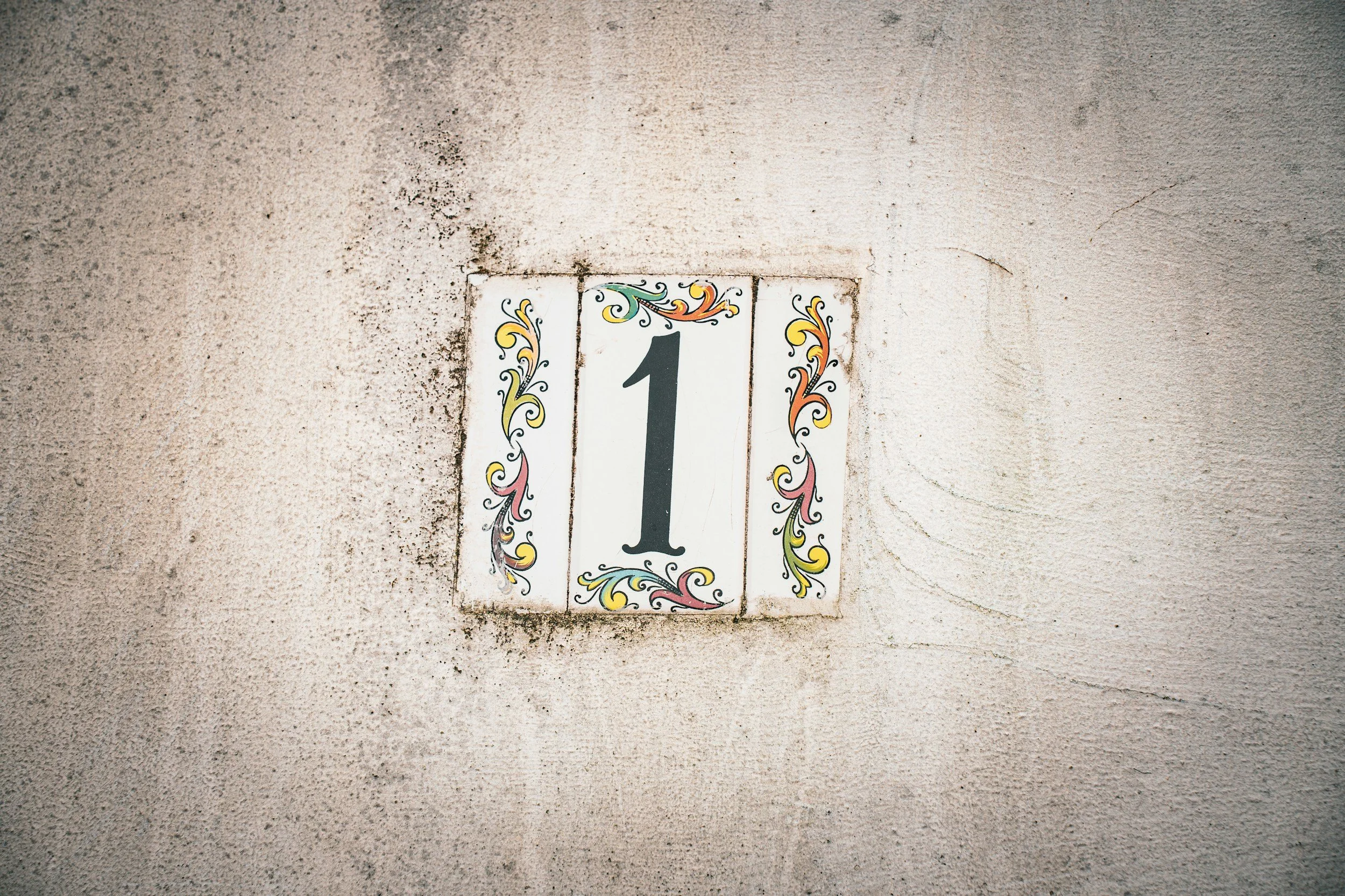 A small wall plaque with decorative floral designs around a number 1, mounted on a rough, beige wall.