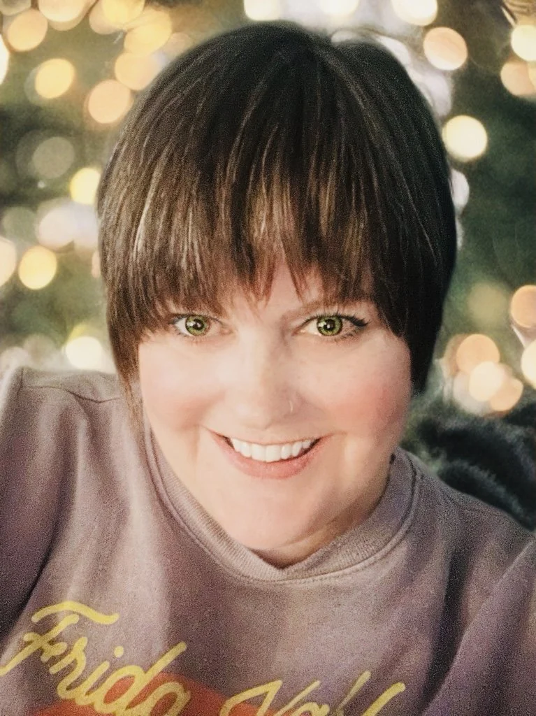 A woman with short brown hair and green eyes smiling in front of a background with blurred yellow and white lights.