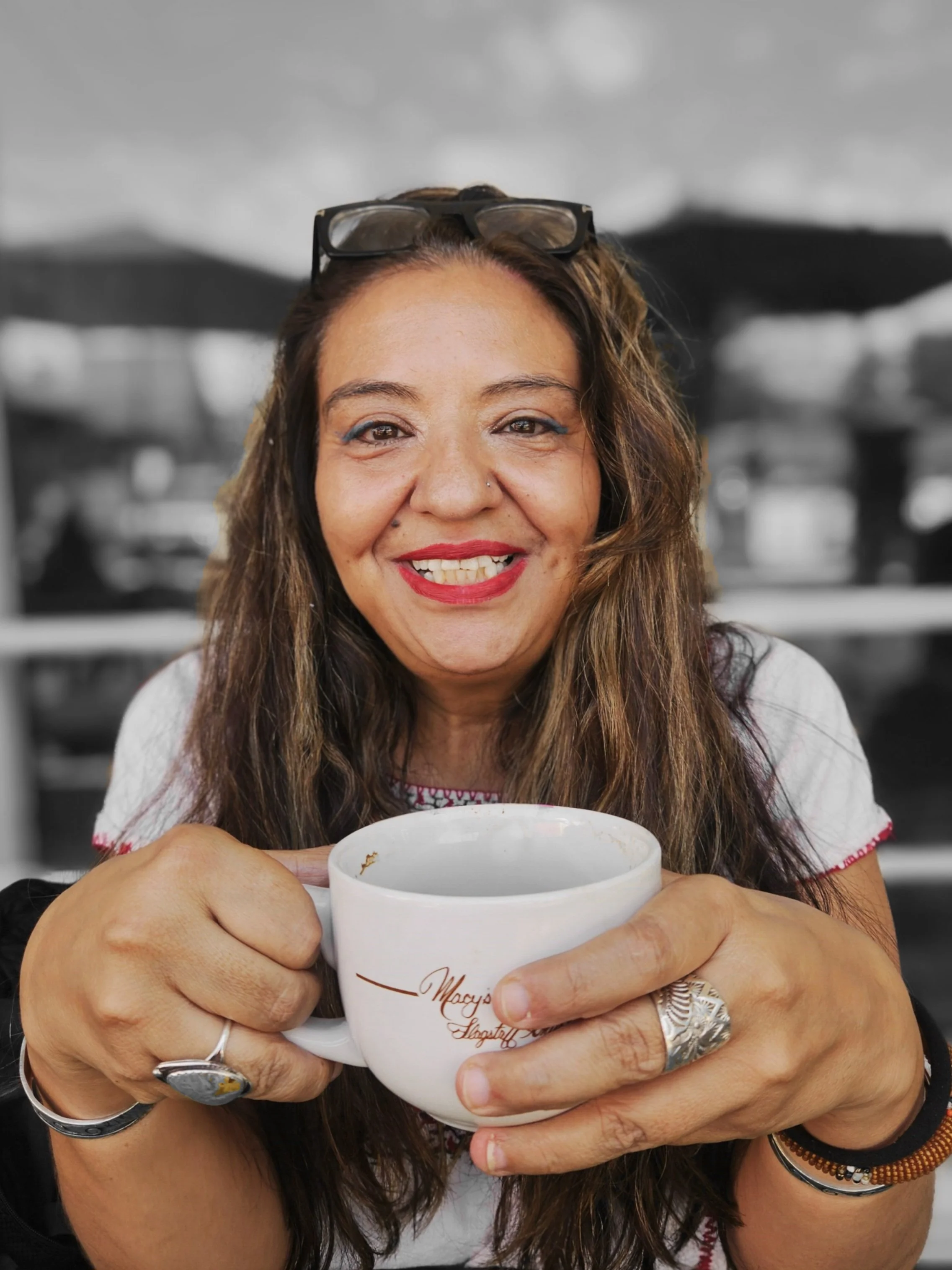 Jennie Marie with long brown hair, red lipstick, and glasses on her head smiling and holding a white coffee mug outdoors.