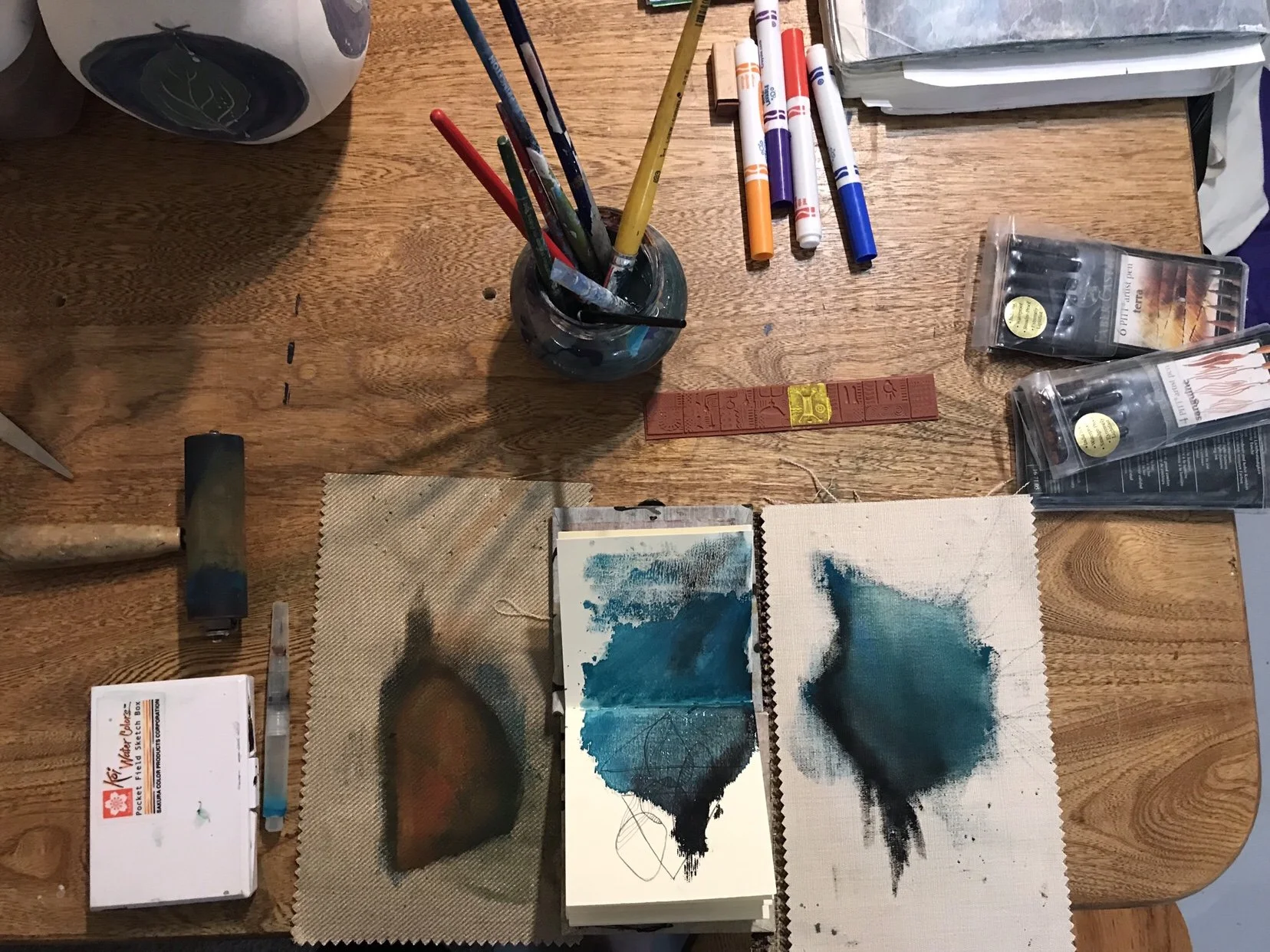 Art supplies on a wooden table, including paintbrushes in a glass jar, markers, a red rubber stamp, paint swatches or artwork, paint sets, and paper with abstract blue and black watercolor paintings.