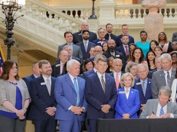 A large group of people, including men and women of various ages, gathered on a staircase inside a building with ornate architectural details. Some individuals are standing and some are sitting, with one person seated at a table in the foreground. Most are dressed in formal business attire, and some are wearing name badges.