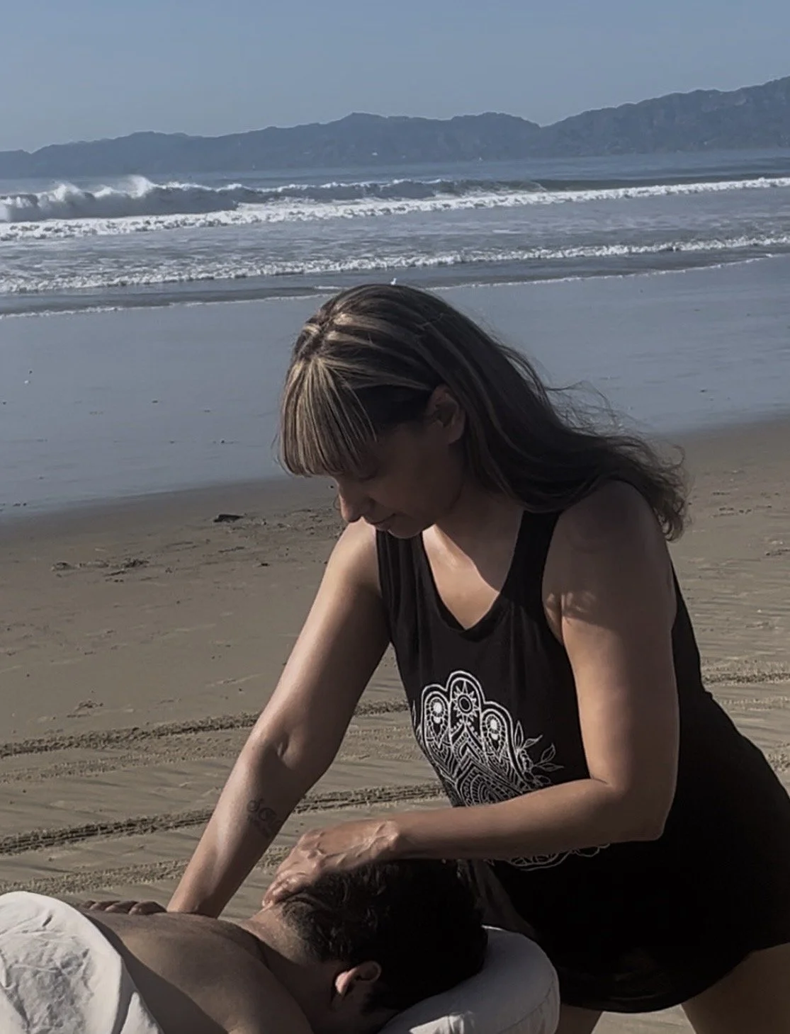 A woman giving a massage on a beach during daytime with ocean waves and mountains in the background.