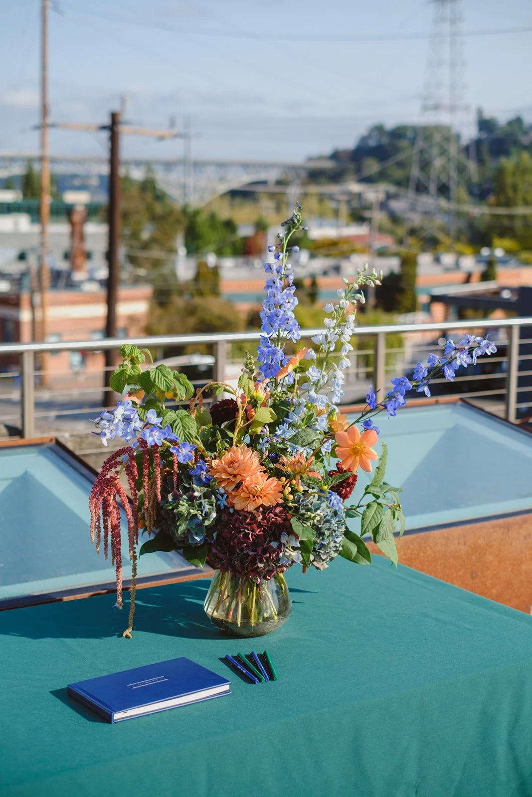 A colorful flower bouquet with blue, orange, and burgundy flowers on a green table, with a blue notebook and pens, on a balcony with railings and a cityscape in the background.