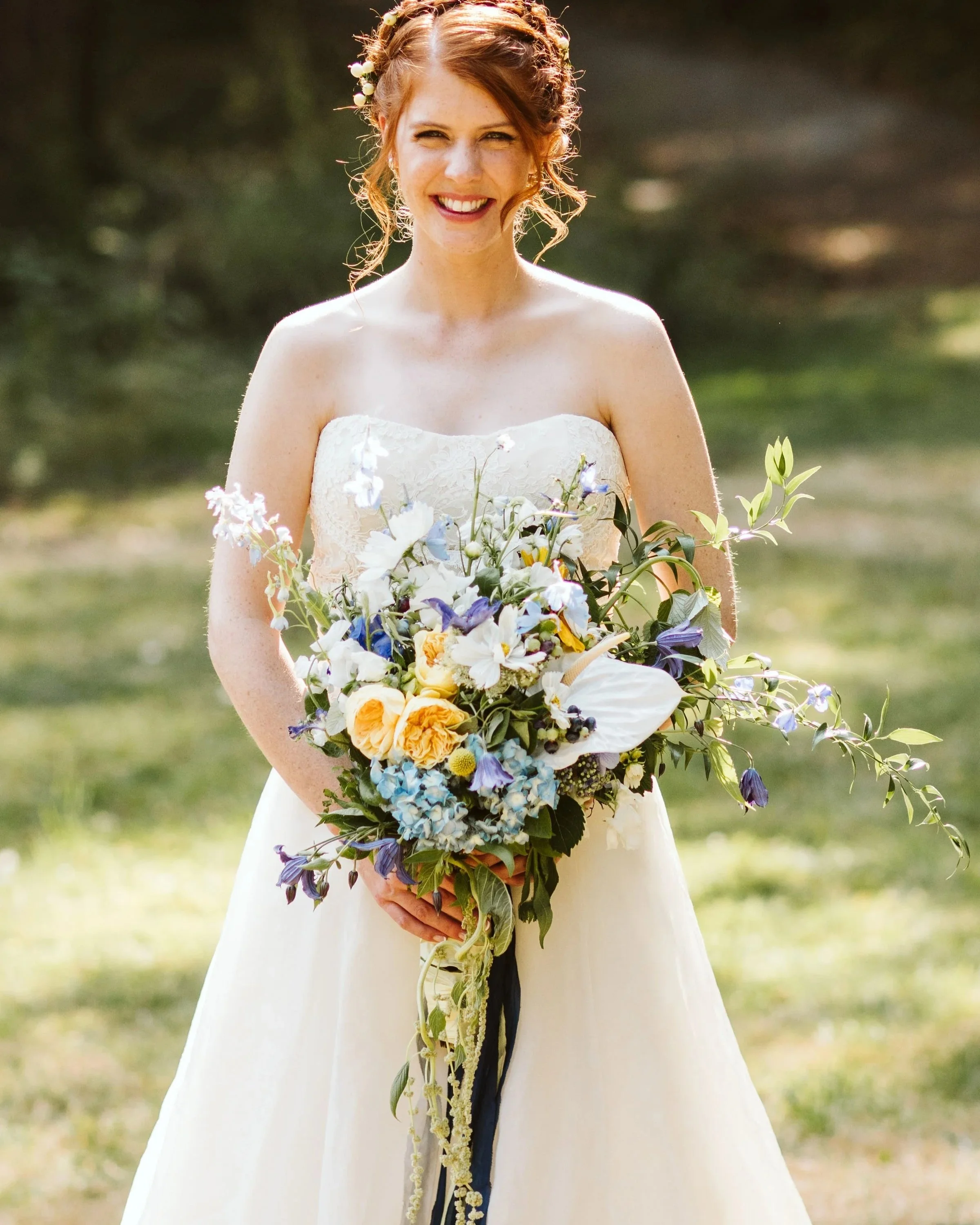A smiling woman in a wedding dress holding a large bouquet of flowers outdoors.