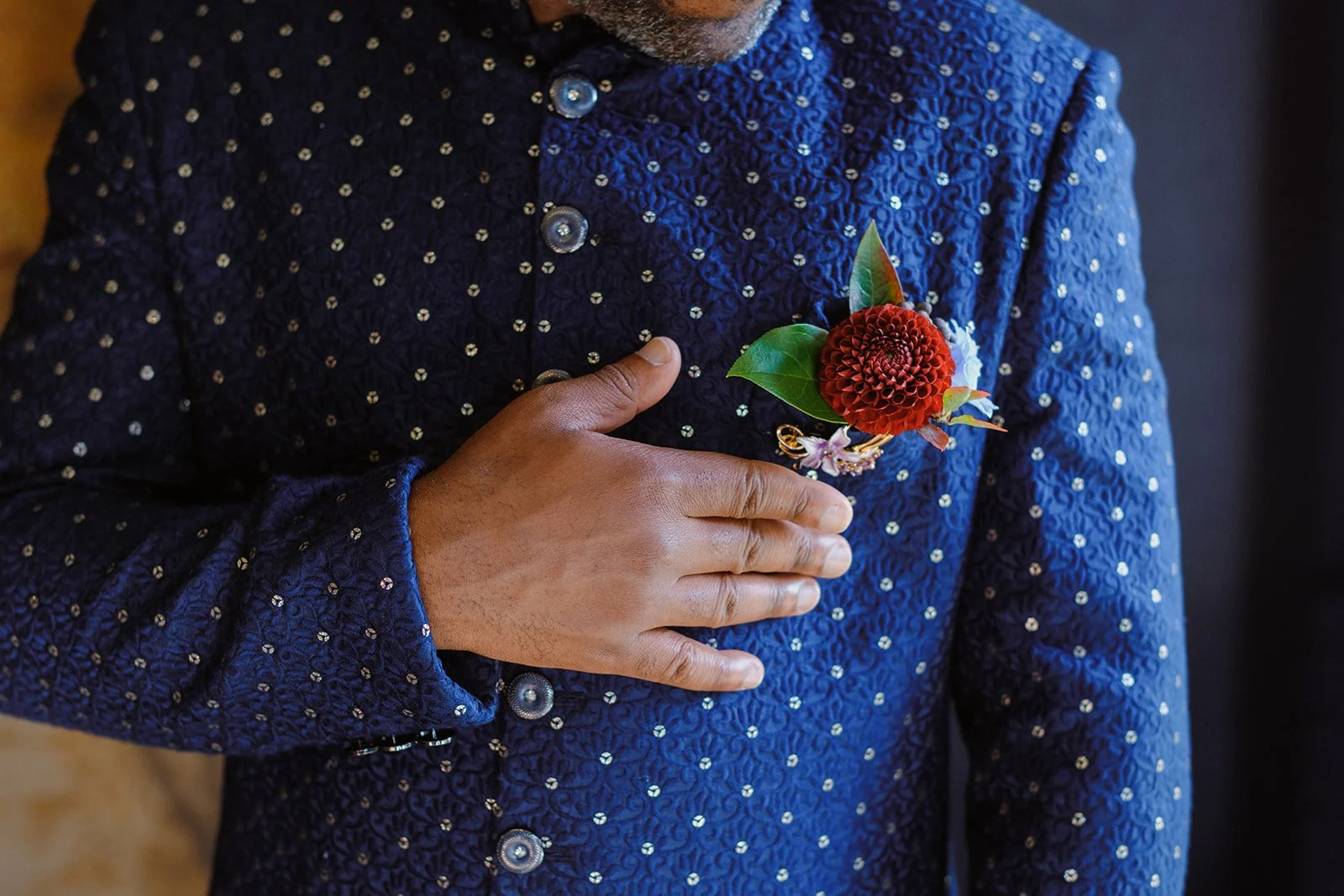 A person wearing a blue textured jacket with silver buttons, holding their hand over their chest, with a red flower boutonniere and green leaves pinned to the jacket.