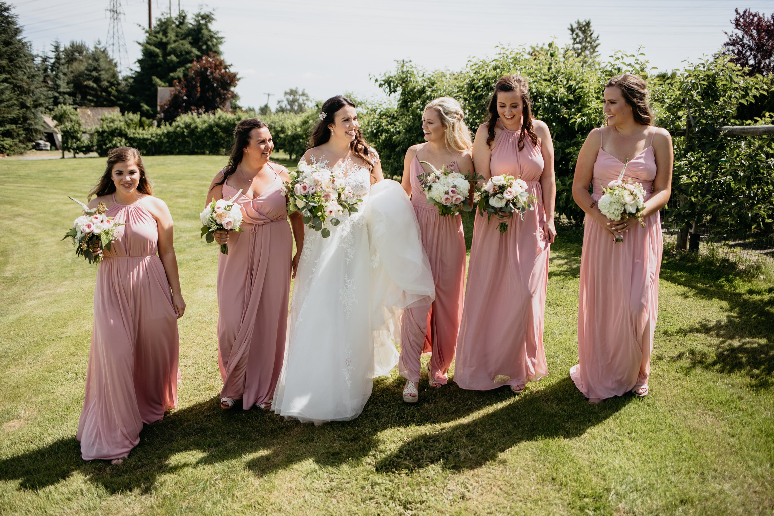A bride and five bridesmaids walking together in a garden. The bride is in a white wedding dress, and the bridesmaids are in pink dresses, all holding bouquets of flowers. They are smiling and enjoying the moment on a sunny day.