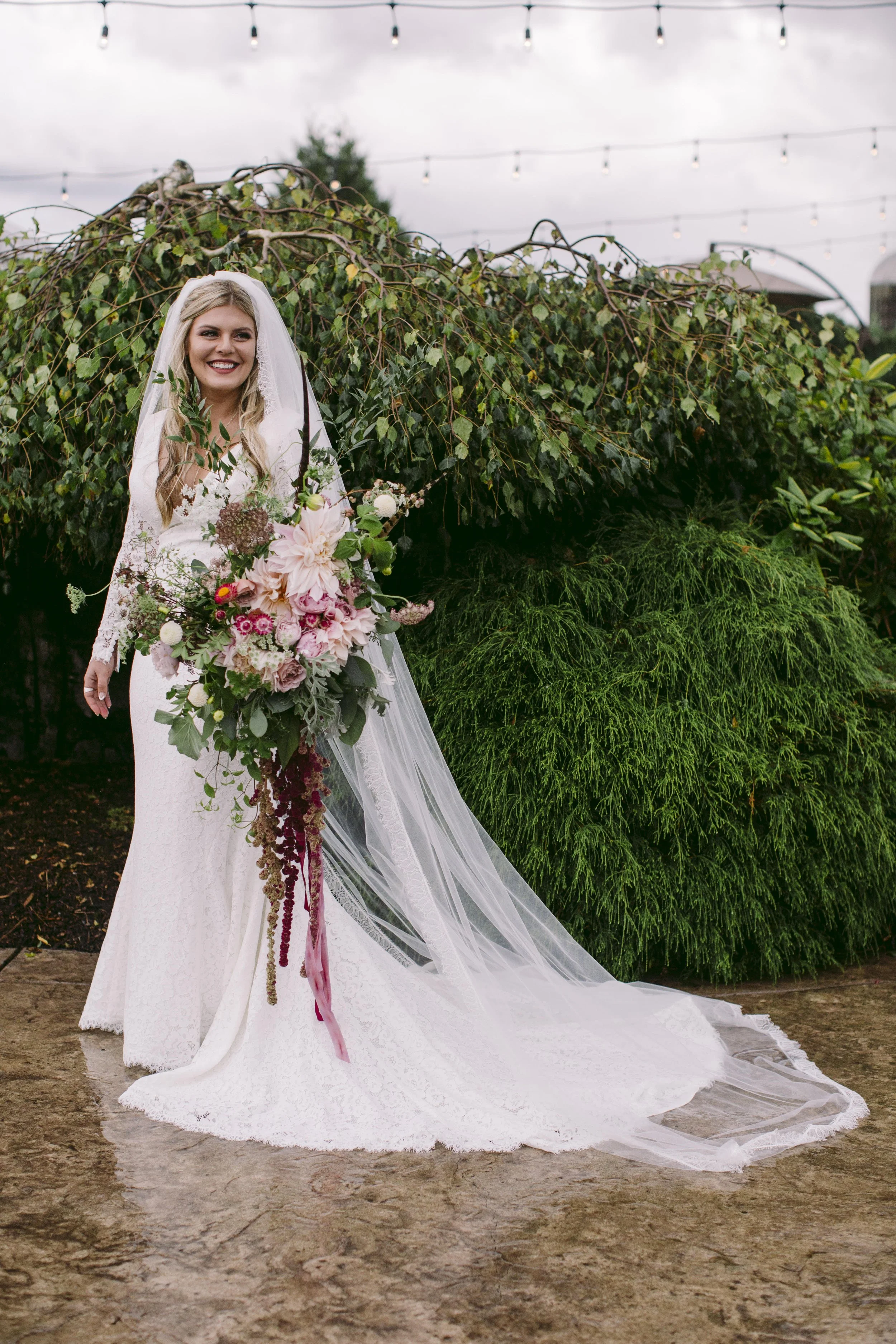 A bride in a white wedding gown and veil holding a large bouquet of pink and white flowers, standing outdoors on a dirt path with green foliage and string lights overhead.