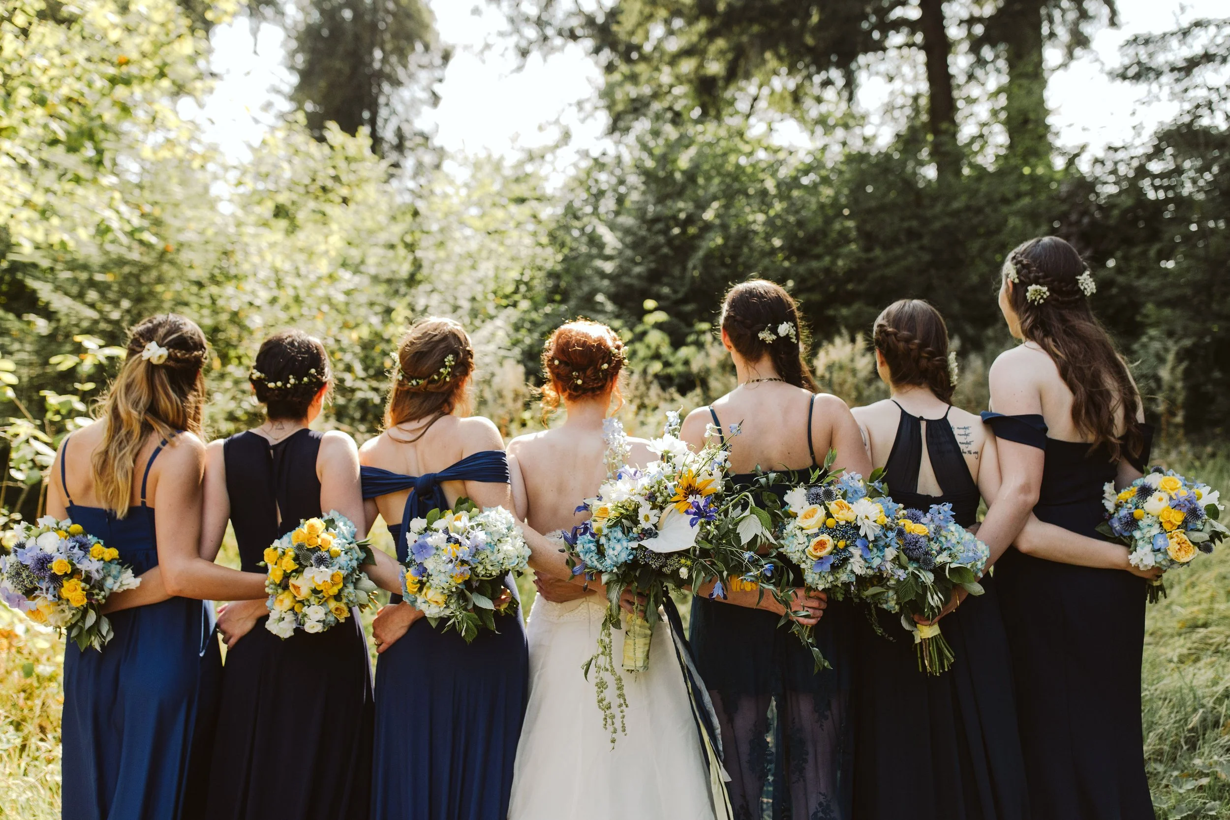 A group of women standing outdoors in a line, dressed in dark blue dresses, holding bouquets of yellow, white, and purple flowers, with their backs to the camera. They have flowers in their hair and are standing in front of a lush, green wooded backg