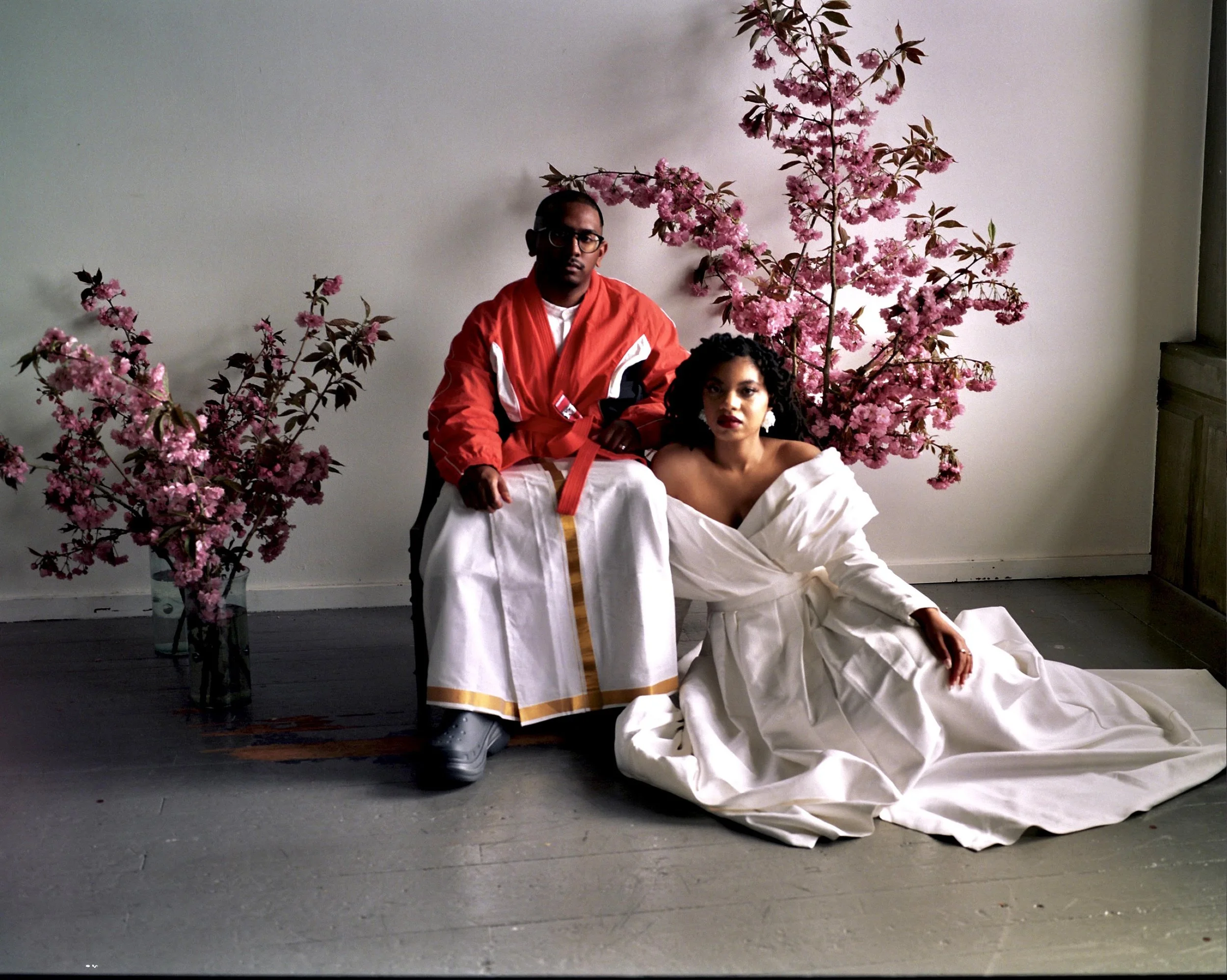 A man and woman pose for a fashion photo amid pink cherry blossom branches in an indoor setting. The man is wearing a white traditional garment with an orange and gold sash, gray shoes, and glasses. The woman is seated on the floor, wearing a white o