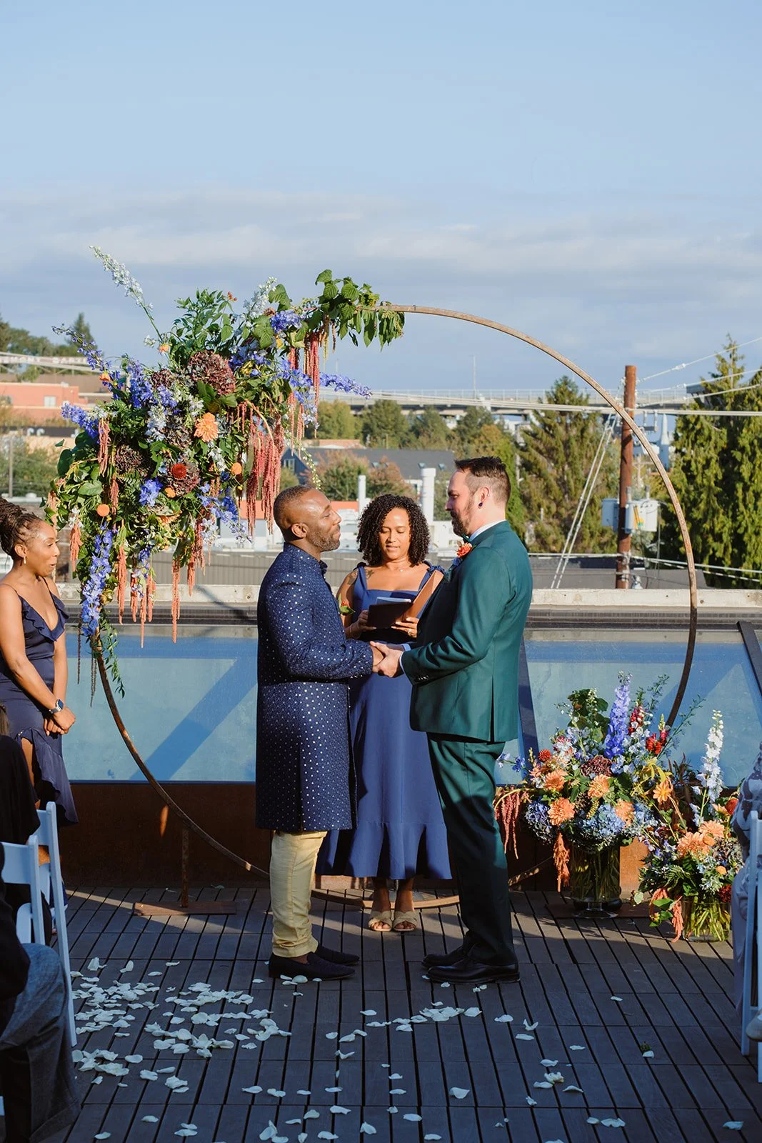 A same-sex wedding ceremony taking place outdoors with a large floral arch. Two men are holding hands and exchanging vows, with a officiant standing behind them. Two women stand on the side watching. There are rose petals on the wooden deck, and the 