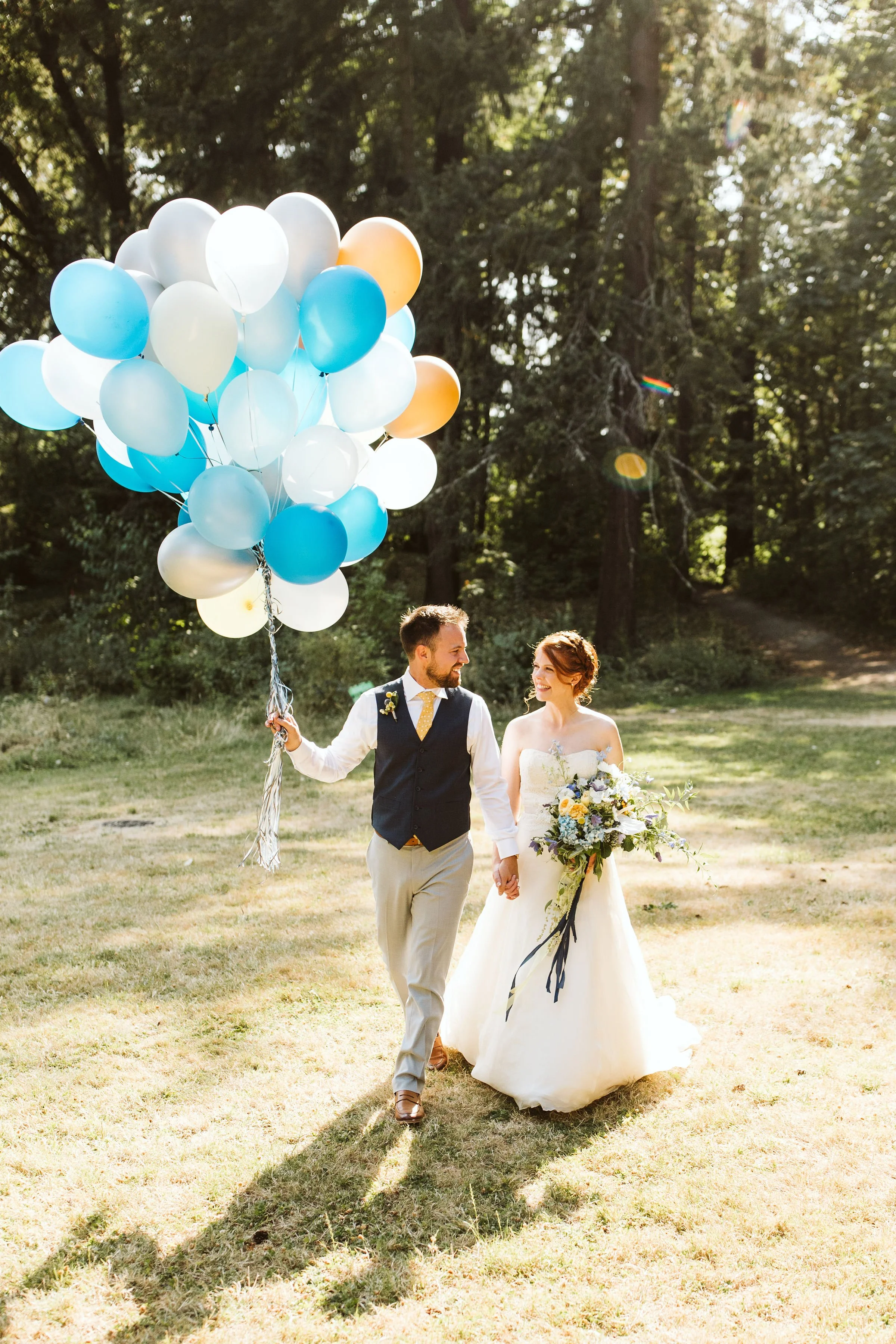 A bride and groom walking together outdoors on a sunny day, holding hands and smiling, with a large bunch of helium balloons in white, blue, and orange in the background.