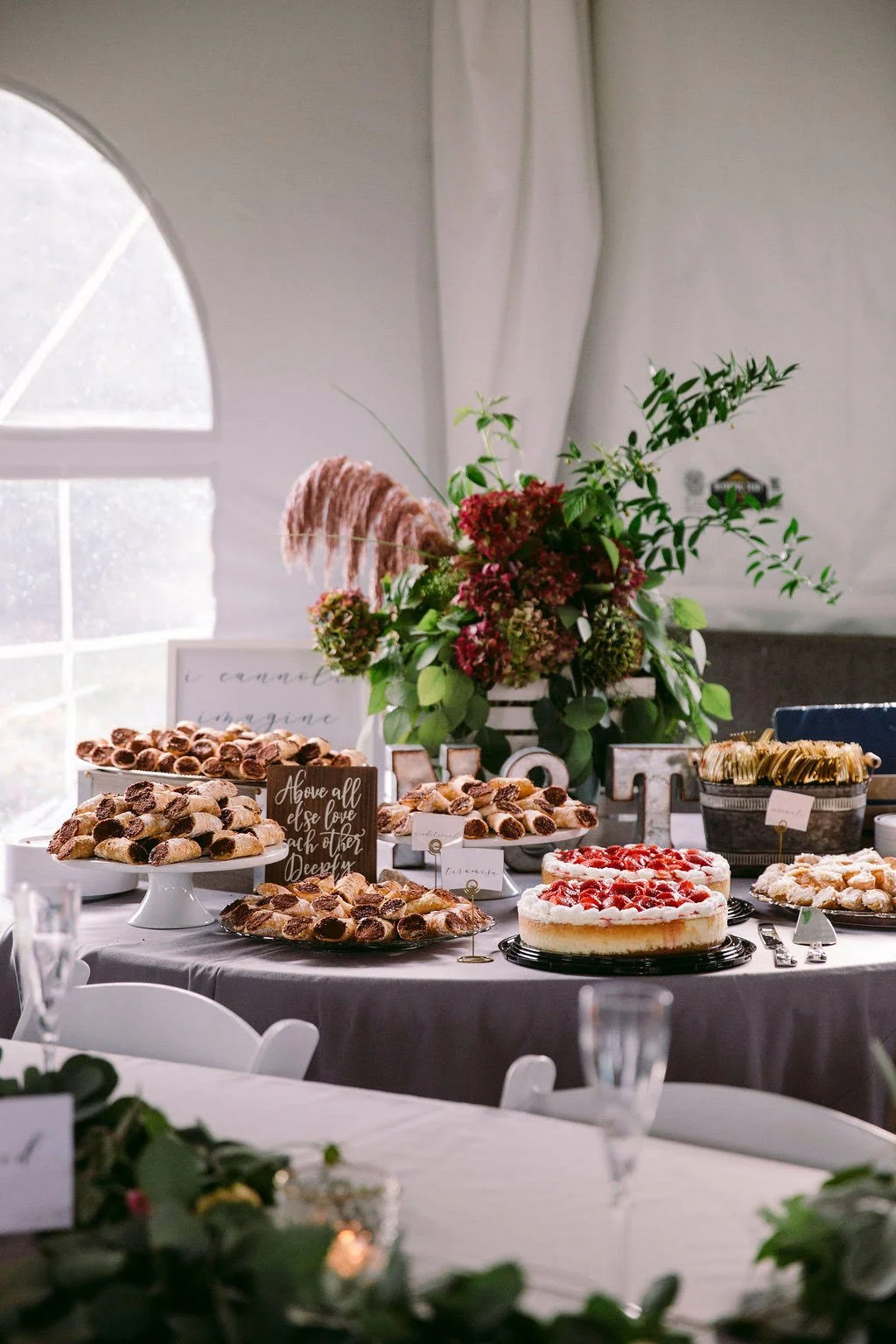 A dessert table with various pastries, a cake topped with strawberries, and a floral centerpiece under a white tent at a celebration or event.