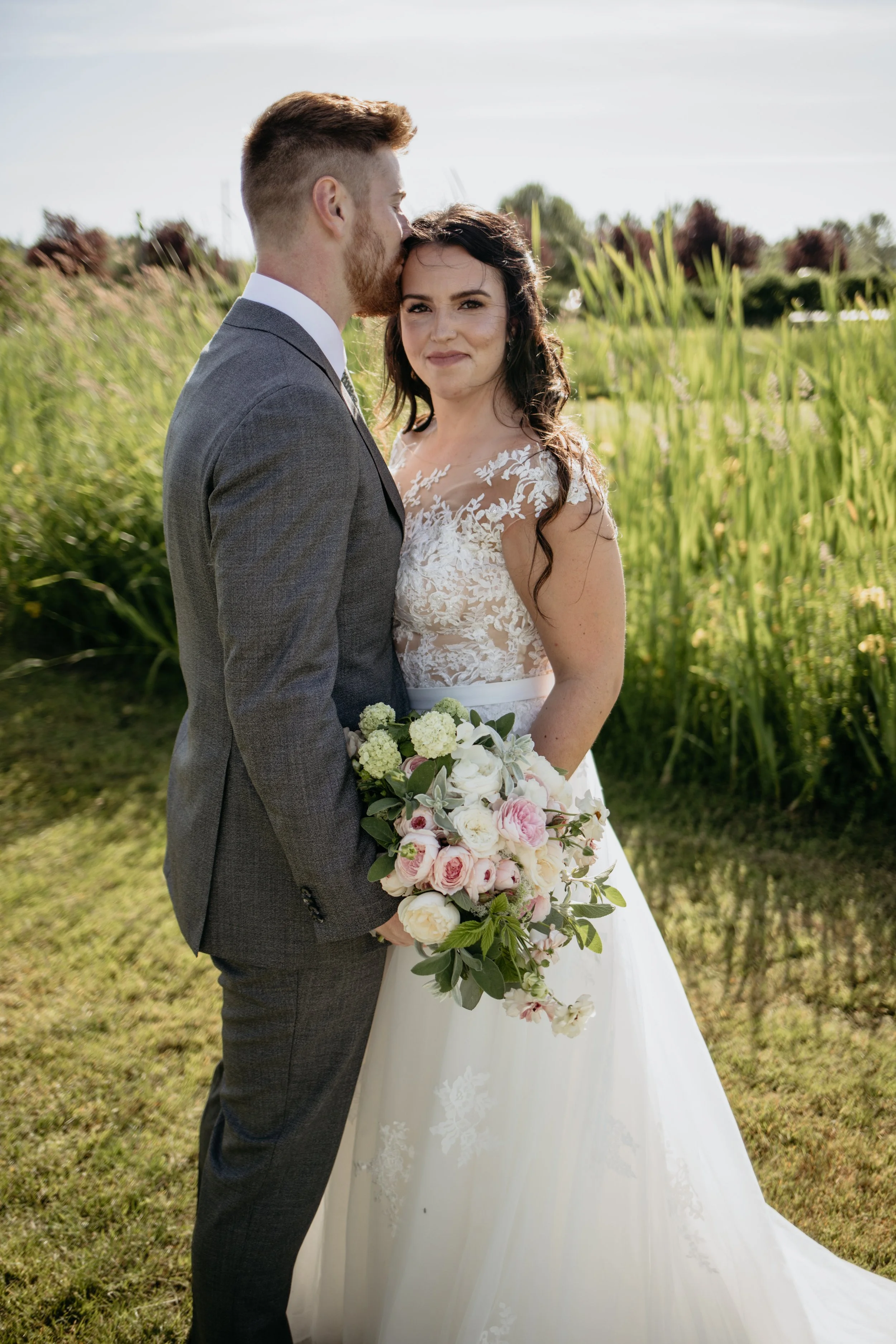 A newlywed couple standing outdoors in a grassy area with tall plants and trees in the background. The groom, with short brown hair and a beard, is wearing a gray suit and is kissing the bride on her forehead. The bride, with long dark hair, is weari