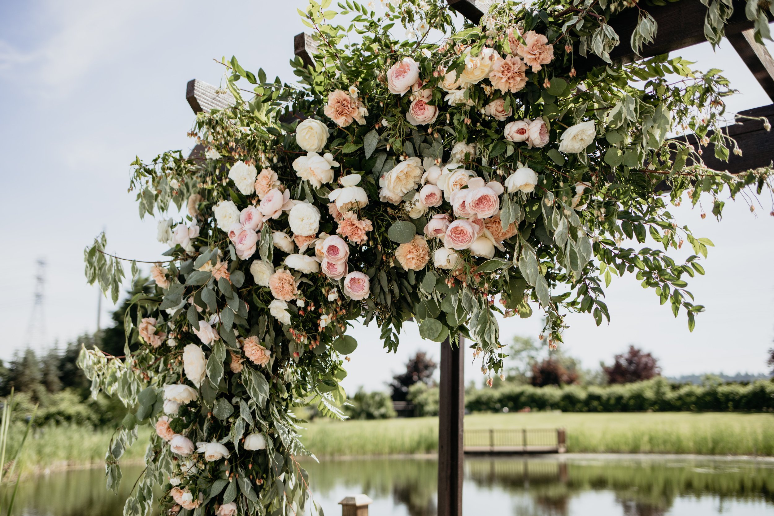 A floral arrangement of pink and white roses and other flowers on a wooden archway, outdoors near a pond and trees.
