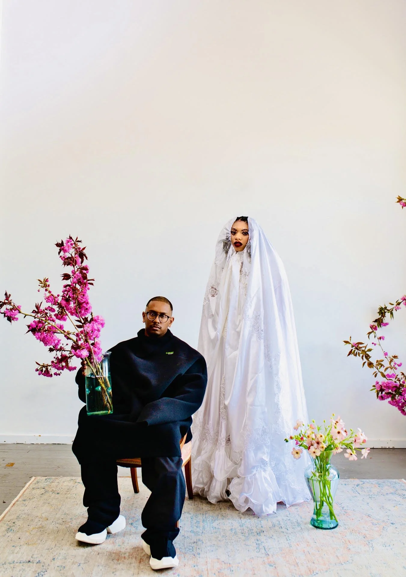 A man sitting on a chair with pink flowers in a vase on his lap, wearing dark clothing and white sneakers, in front of a woman dressed in a white gown, with pink and purple flowers surrounding them against a white wall background.