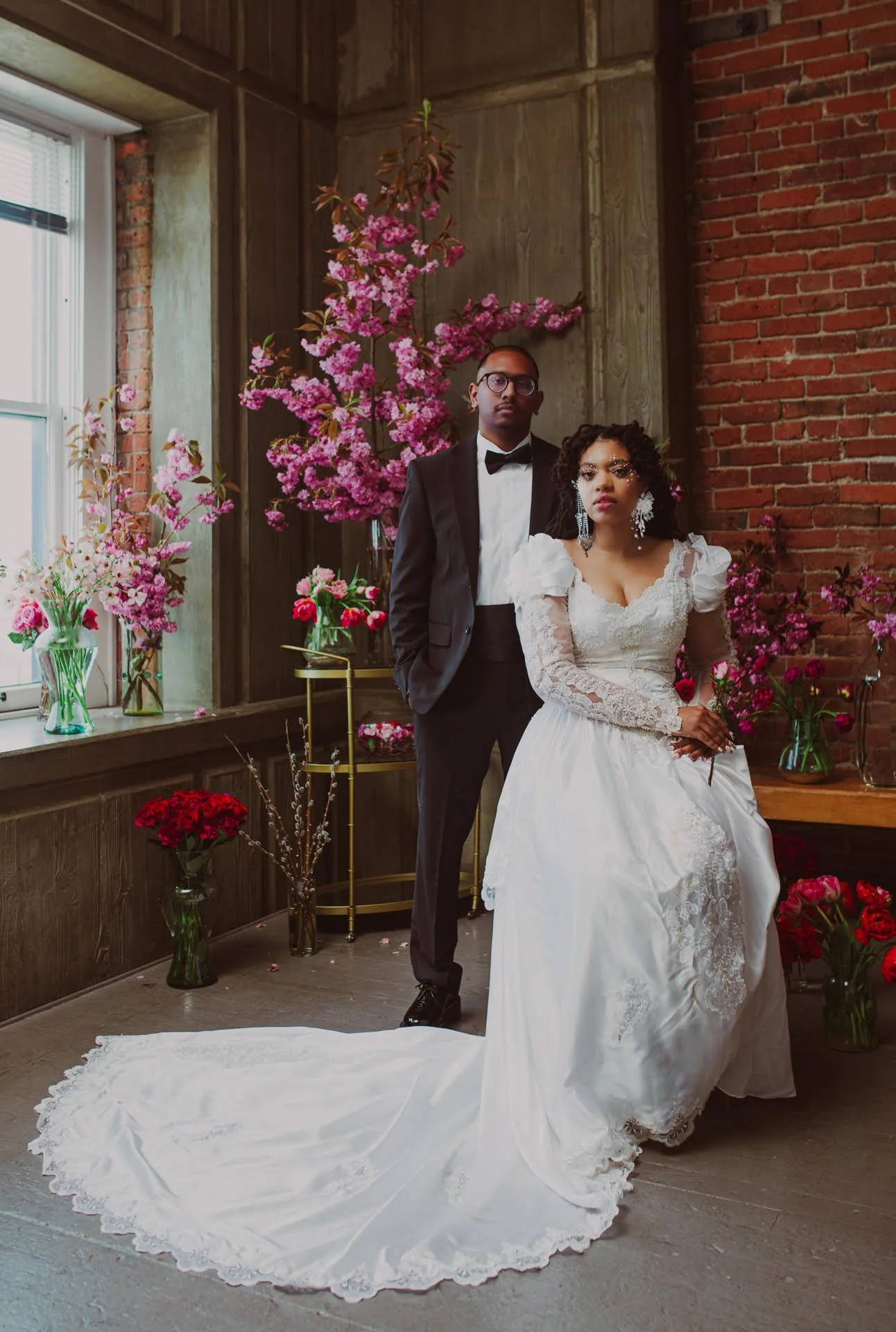 A bride in a white wedding dress with lace details and a long train, and a groom in a black tuxedo with a bow tie, standing indoors with pink and red floral arrangements and a brick wall in the background.