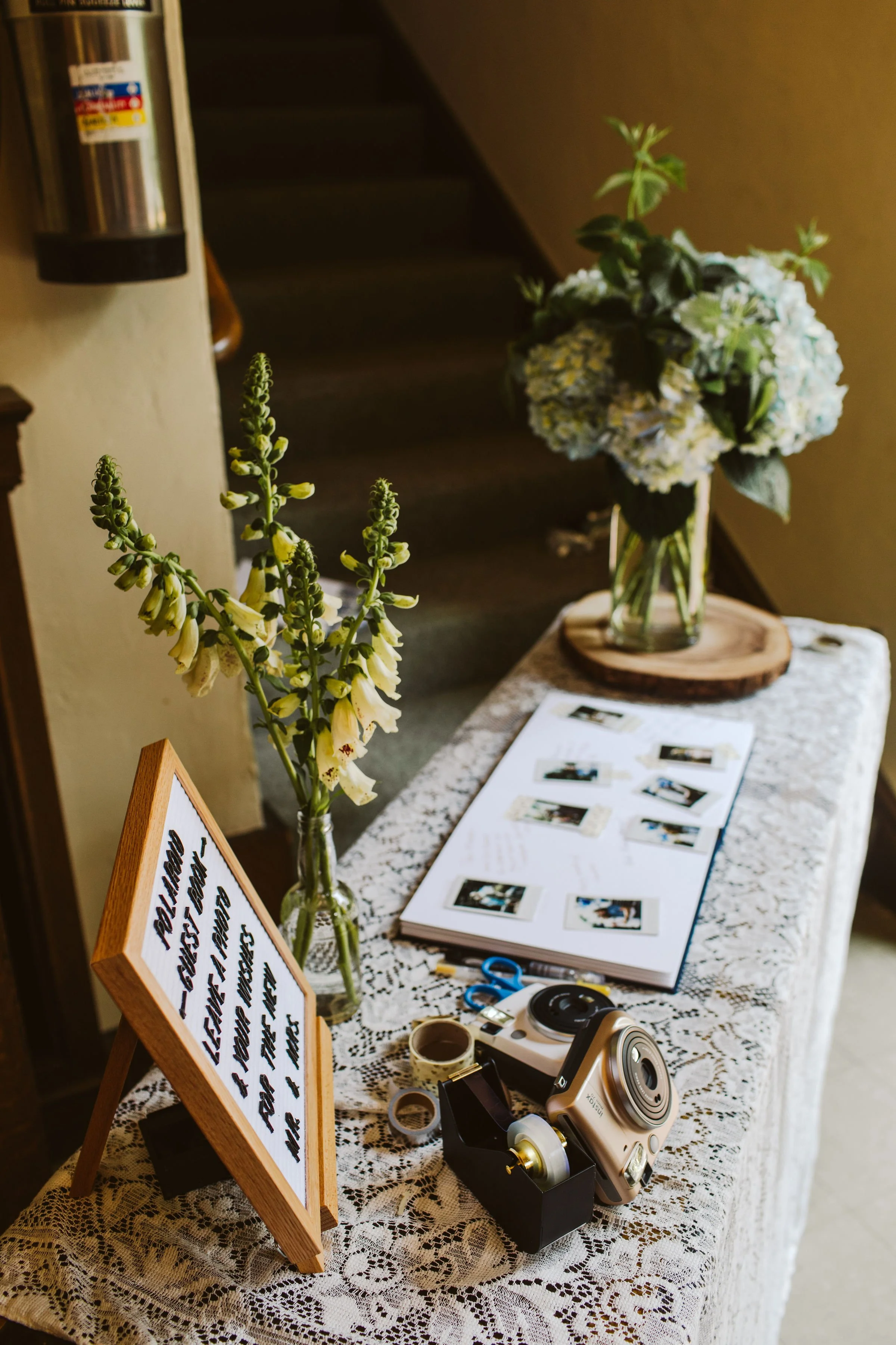 Table decorated with lace tablecloth displaying a sign, a scrapbook, tape, a camera, and floral arrangements in vases, along with scissors, tape, and other small items.