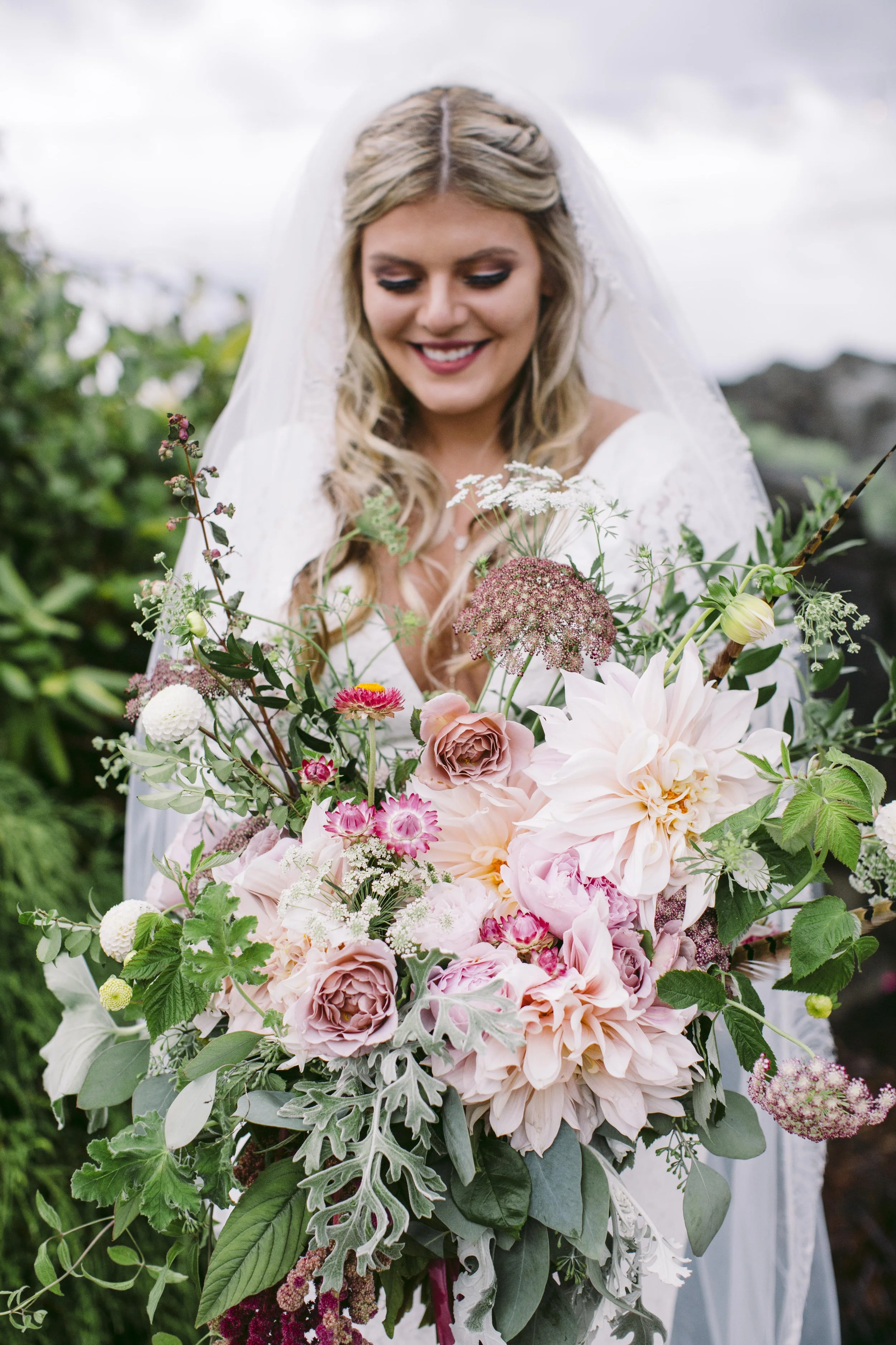 A bride holding a large bouquet of pink and white flowers, with green foliage, outdoors on a cloudy day.