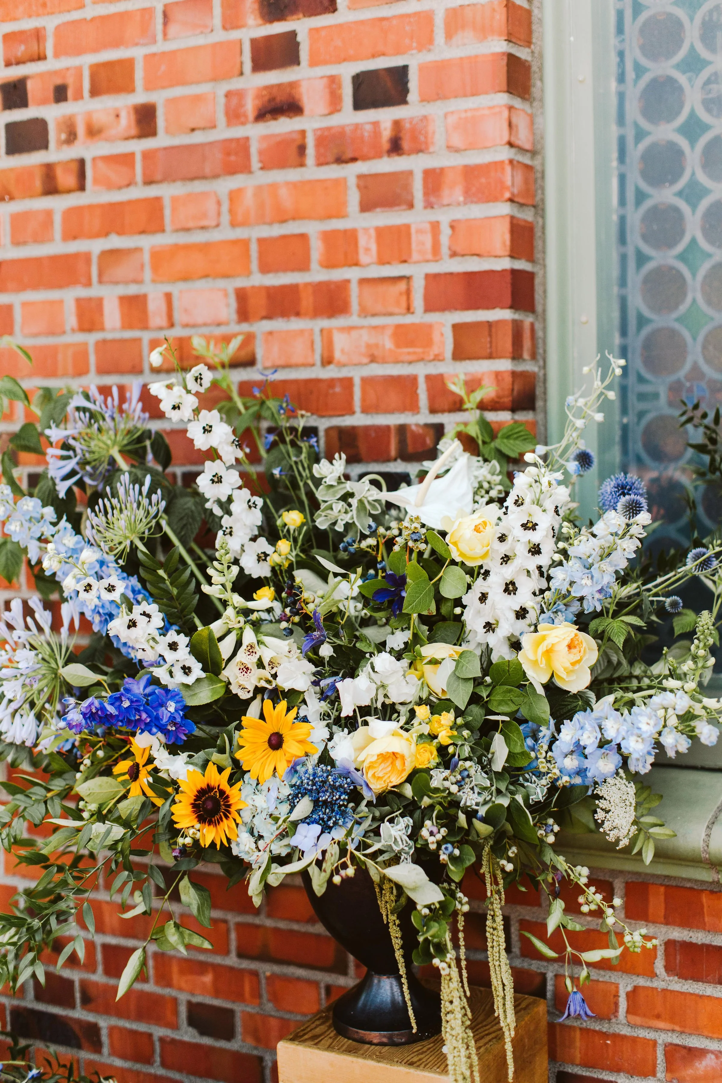 A large colorful flower arrangement in a dark pedestal vase on a wooden table in front of a red brick wall with a metal and glass window to the side.