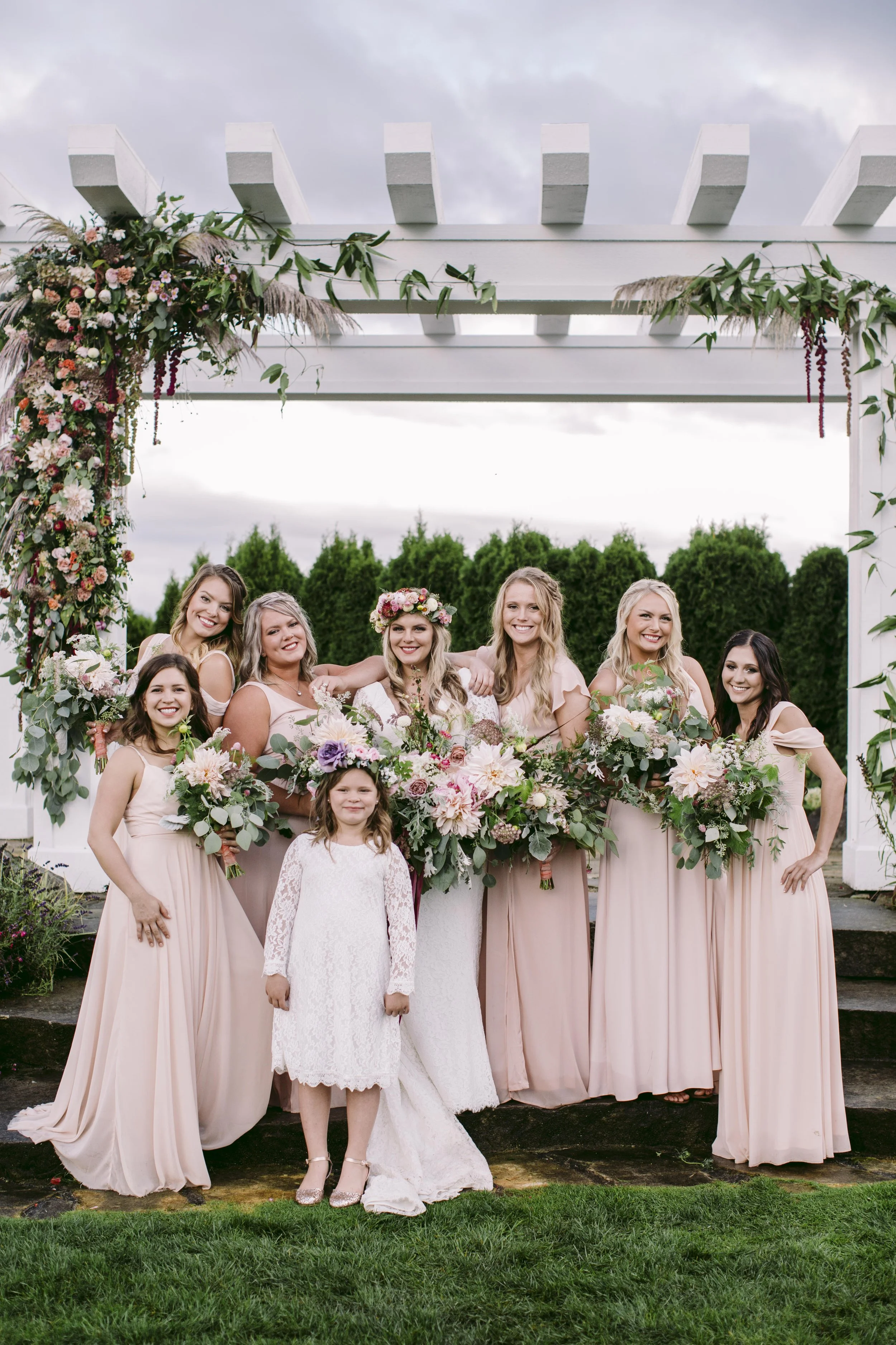 A bride in a white lace dress standing among bridesmaids in blush pink dresses at an outdoor wedding. The group poses under a white wooden arch decorated with flowers and greenery, with a cloudy sky and green trees in the background.