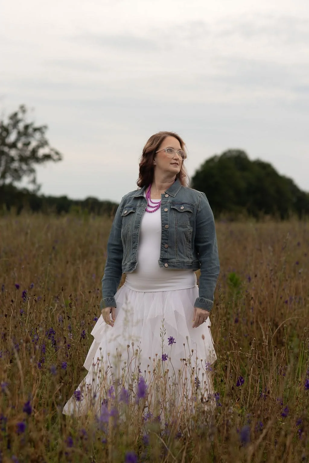 A woman standing in a field of tall grass with purple flowers, wearing a denim jacket, white dress, purple necklace, and glasses, looking to her right with a cloudy sky background.