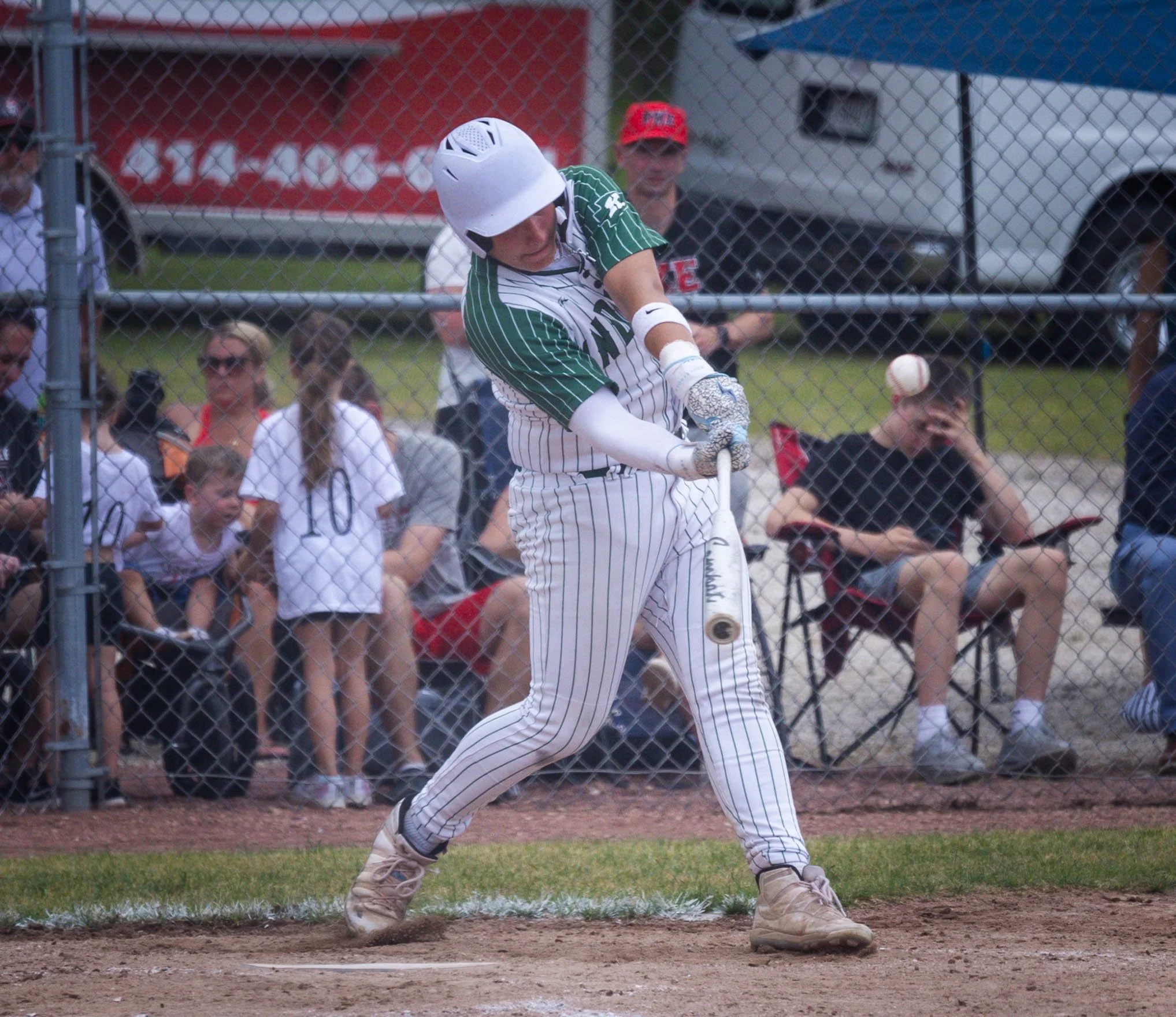 A baseball player swinging at a pitched ball at a baseball field, with spectators watching behind a chain-link fence.