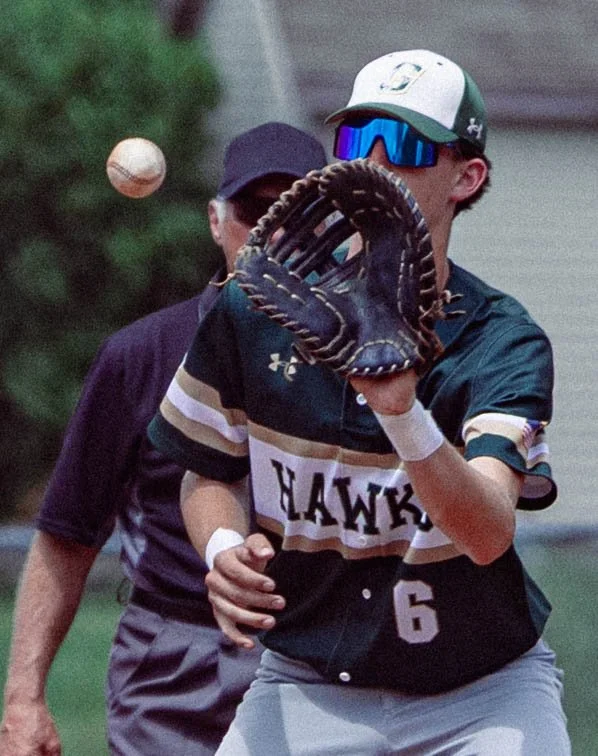 A young baseball player in a black and white uniform with the word "Hawks" and the number 6, wearing sunglasses and a cap, catching a baseball with a glove during a game.