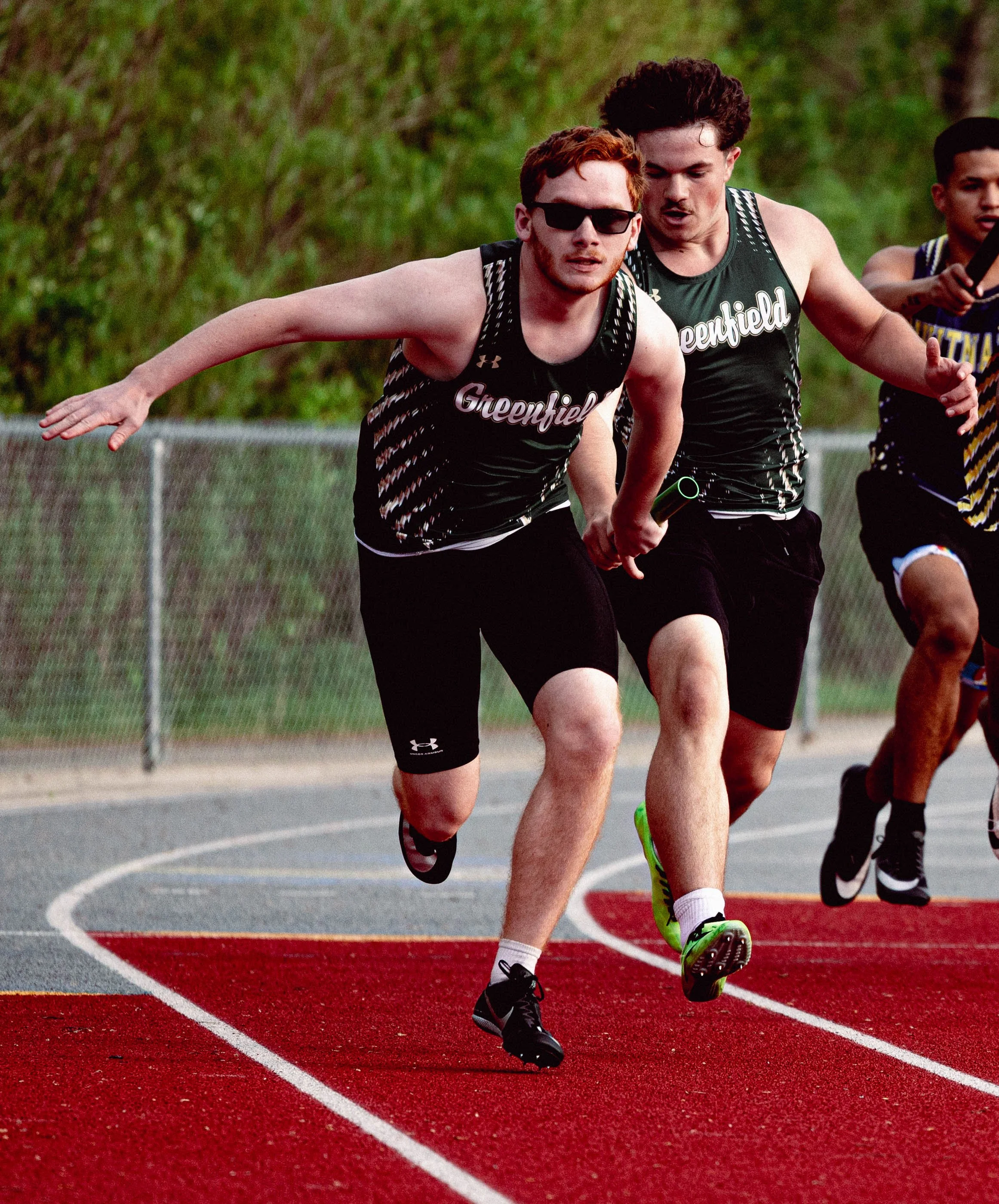 Track athletes running on a red outdoor track with white lane markings, with a green fence and trees in the background.