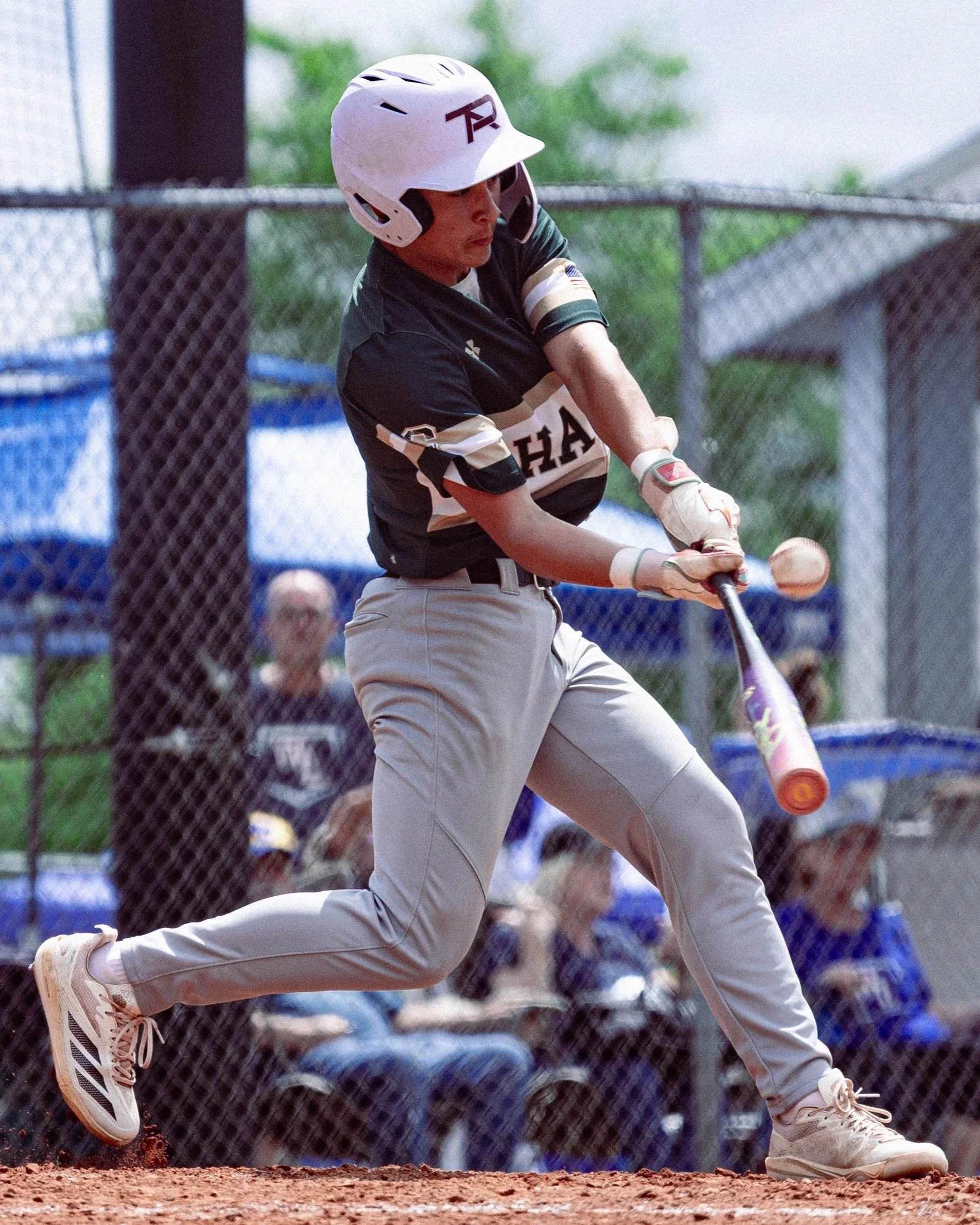 A baseball player is sliding into a base, with only part of his body visible, holding a bat in his right hand, wearing a white helmet, a black and beige jersey, and beige pants, on a baseball field with a chain-link fence and spectators in the backgr