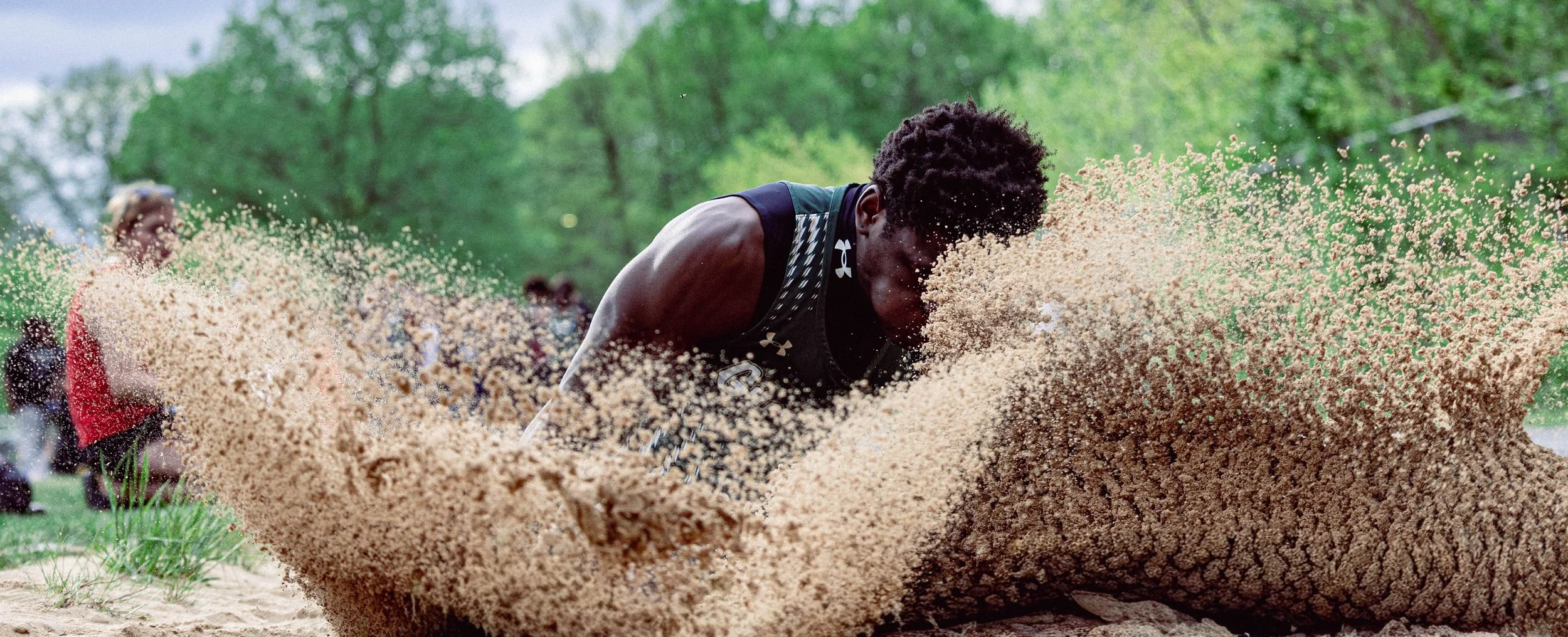 A male athlete in black sportswear with a white logo is diving into a sandpit, creating a cloud of sand, with green trees and spectators in the background.