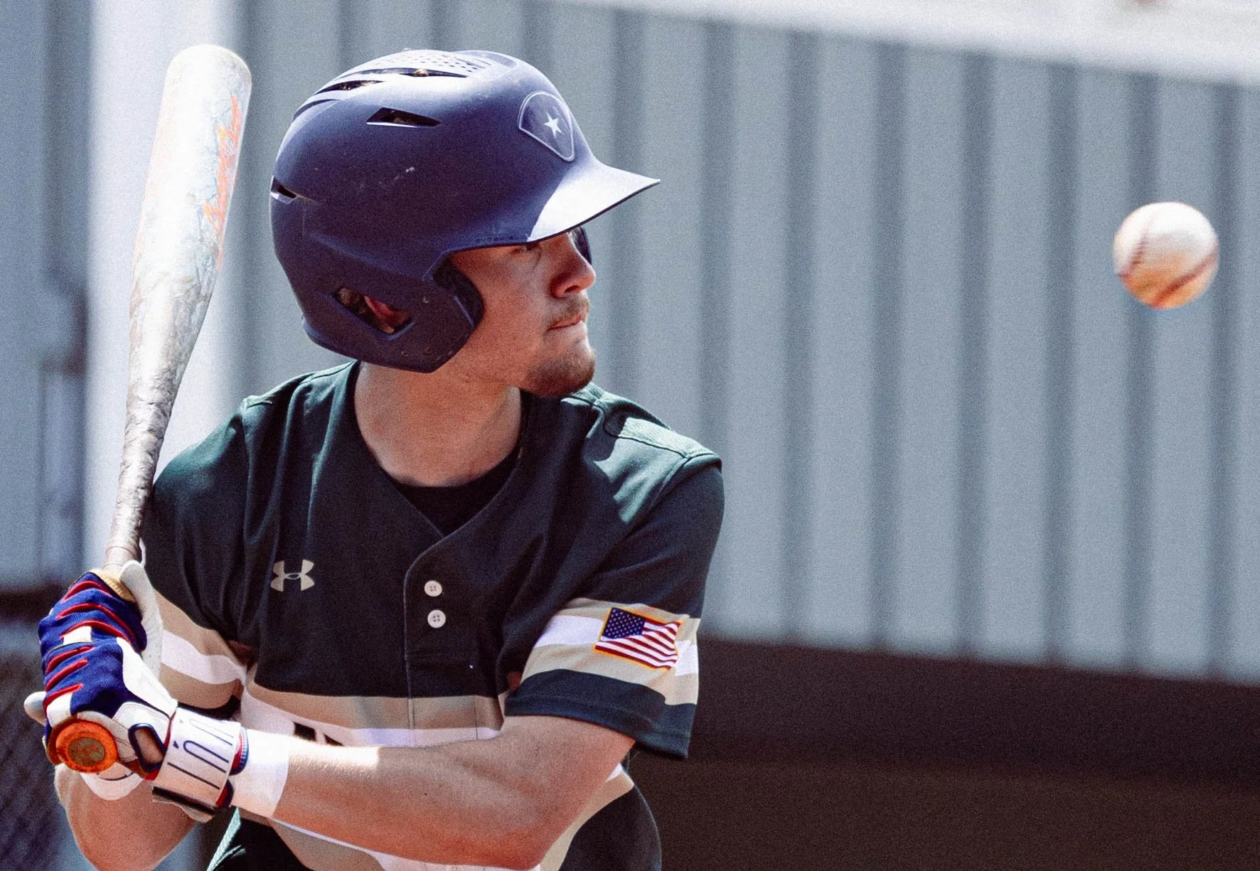 A baseball player wearing a dark helmet with a star logo, a black and striped uniform with an American flag patch, and batting gloves, holding a bat and preparing to hit a baseball in an outdoor setting.