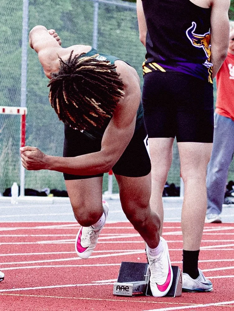 A female athlete with dreadlocks is at the starting position on a running track, ready to sprint, with her front foot on a starting block.