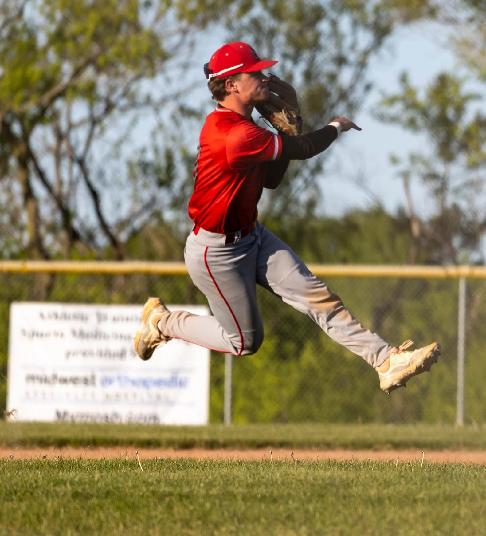 A baseball player in a red jersey and grey pants is jumping in the air to catch a ball with a glove. The background shows a baseball field with a fence and trees.