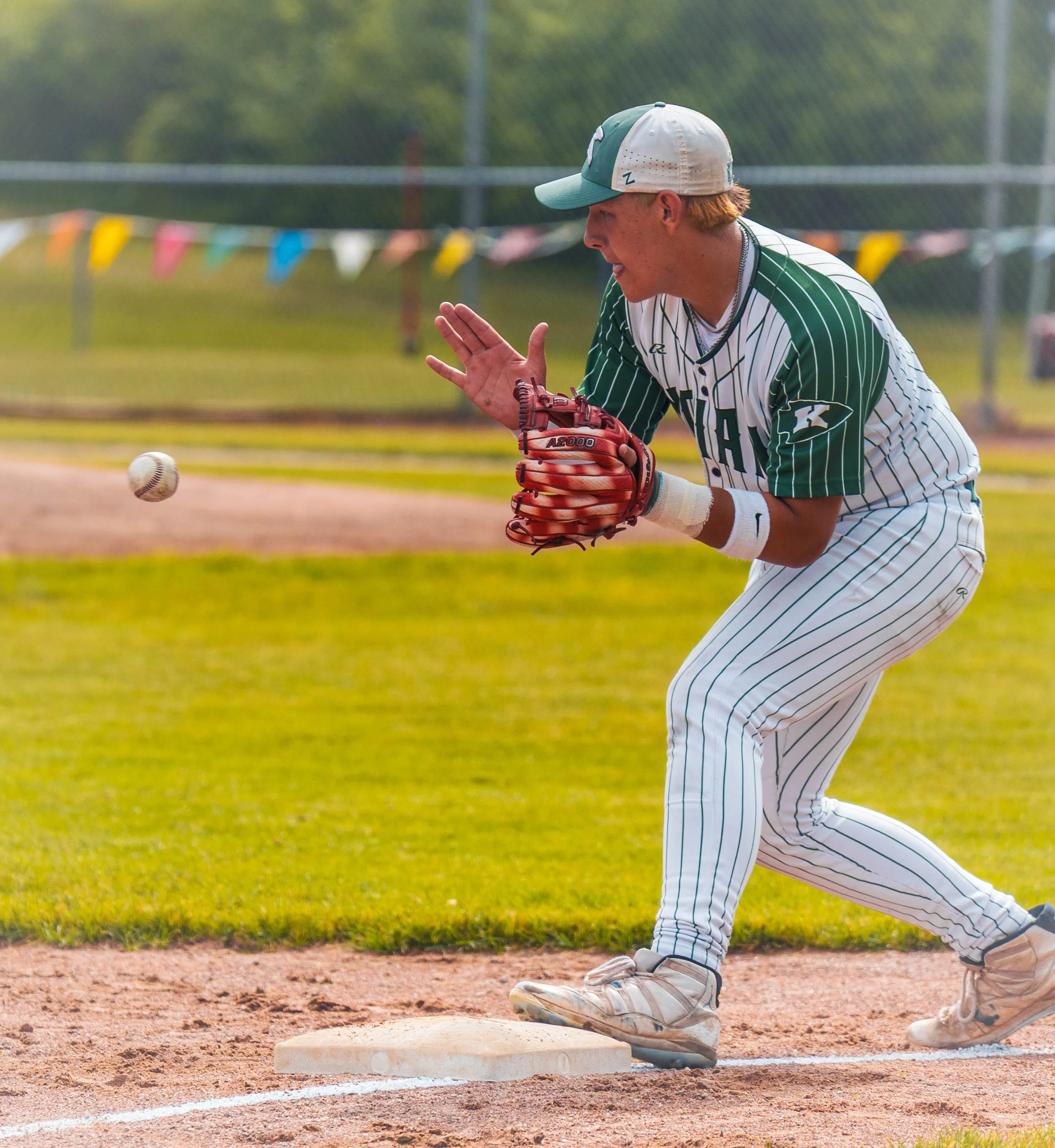 A baseball player in a green and white striped uniform is positioned on a baseball field, preparing to catch a ball with a glove. The player is wearing a cap and has a focused expression. The scene features a grassy background, a chain-link fence, an