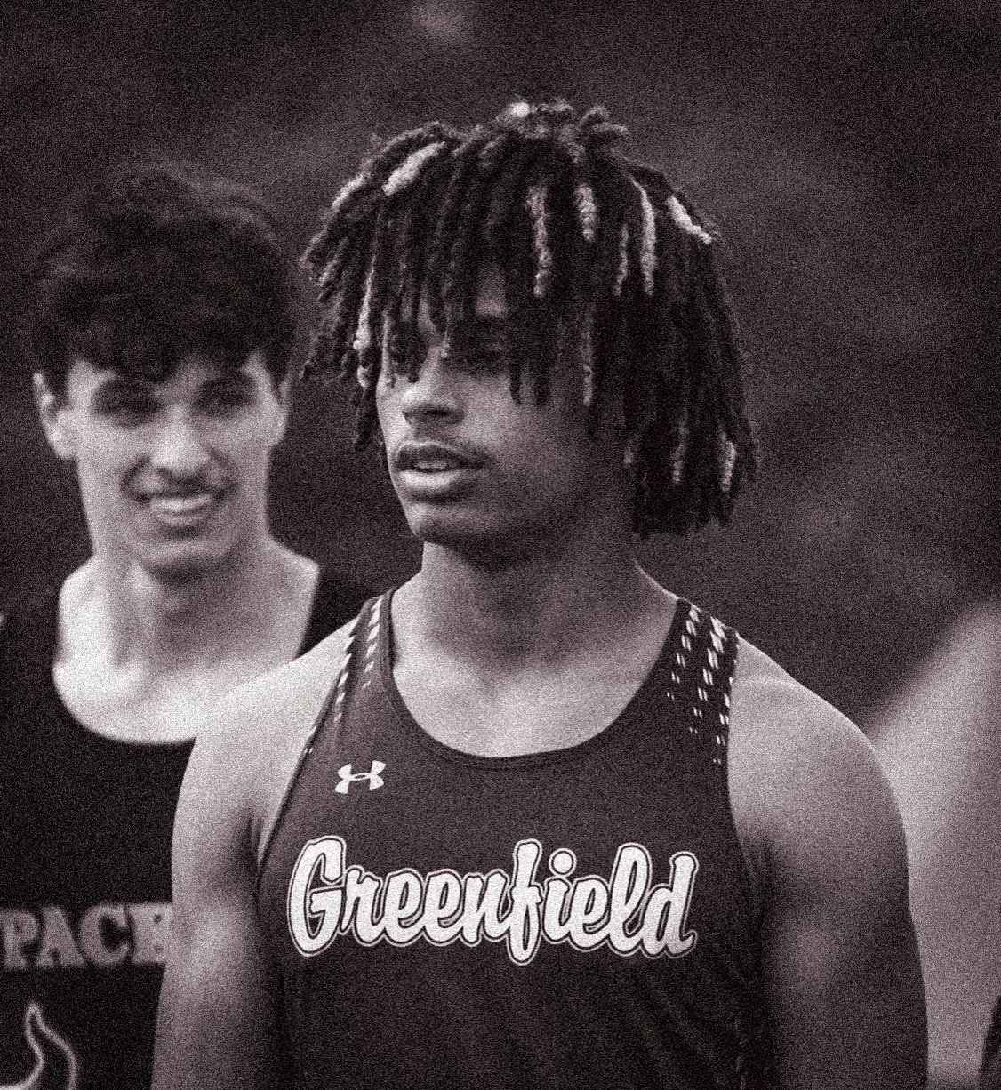 Black and white photo of a young man with dreadlocks wearing a sports jersey with the word "Greenfield" on it, standing outdoors, with a smiling woman with short dark hair behind him.