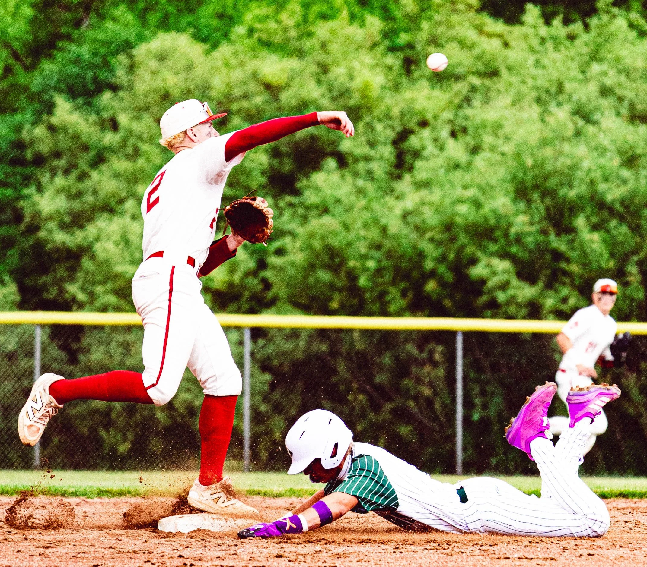 A baseball player in a white and red uniform is jumping to catch a ball in the outfield, while an opposing player in a striped uniform slides into a base, with other players and a fence visible in the background.