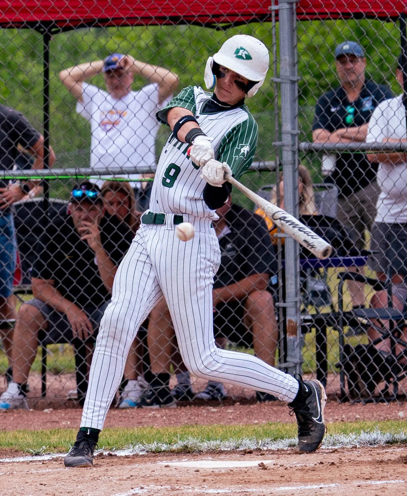 A baseball player swinging a bat during a game, wearing a white uniform with green pinstripes, black eye black, and a helmet. Spectators are watching behind the fence.