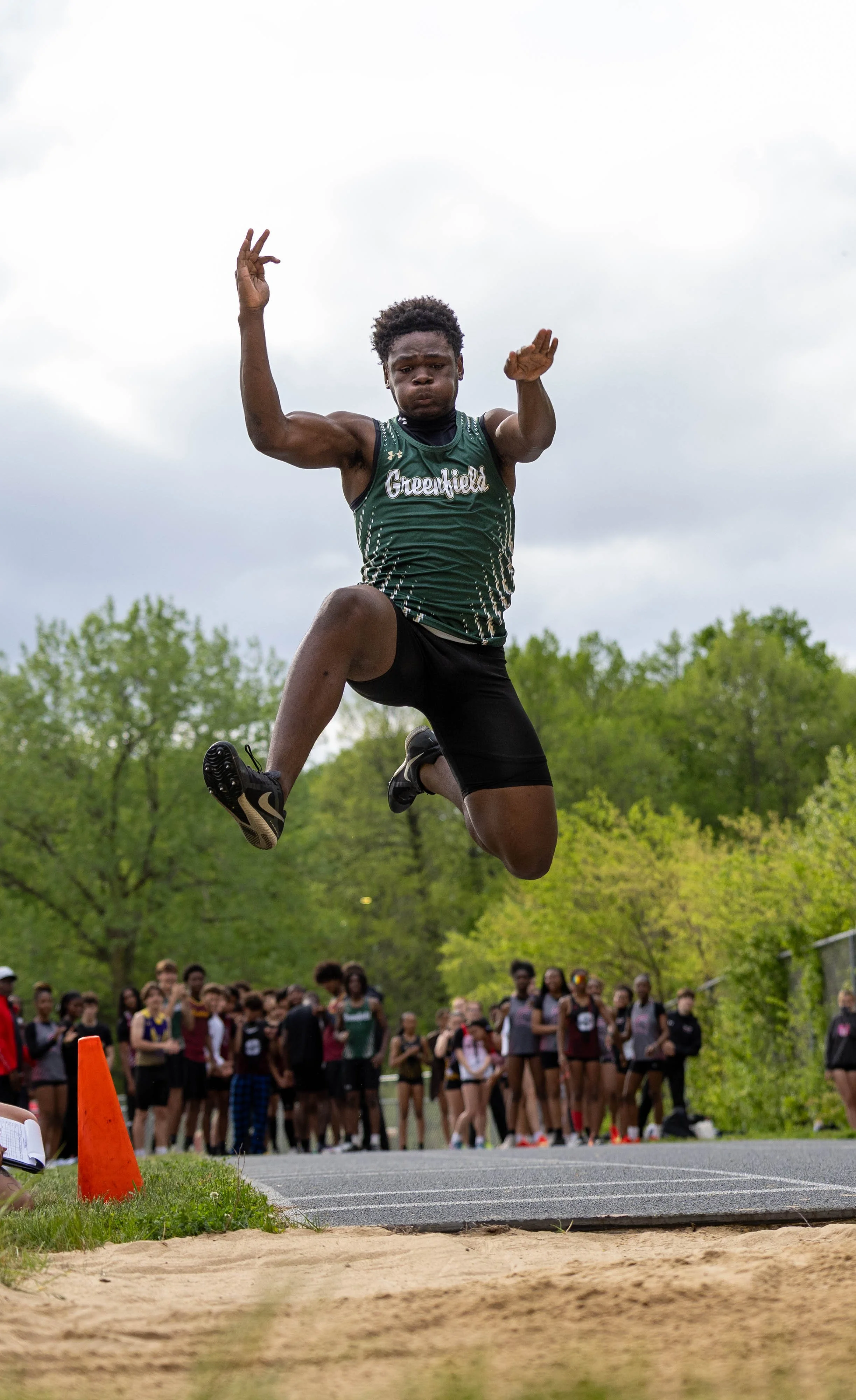 A male long jumper in green athletic uniform mid-air during a jump at a track and field event, with a crowd of spectators and other athletes in the background under cloudy sky.