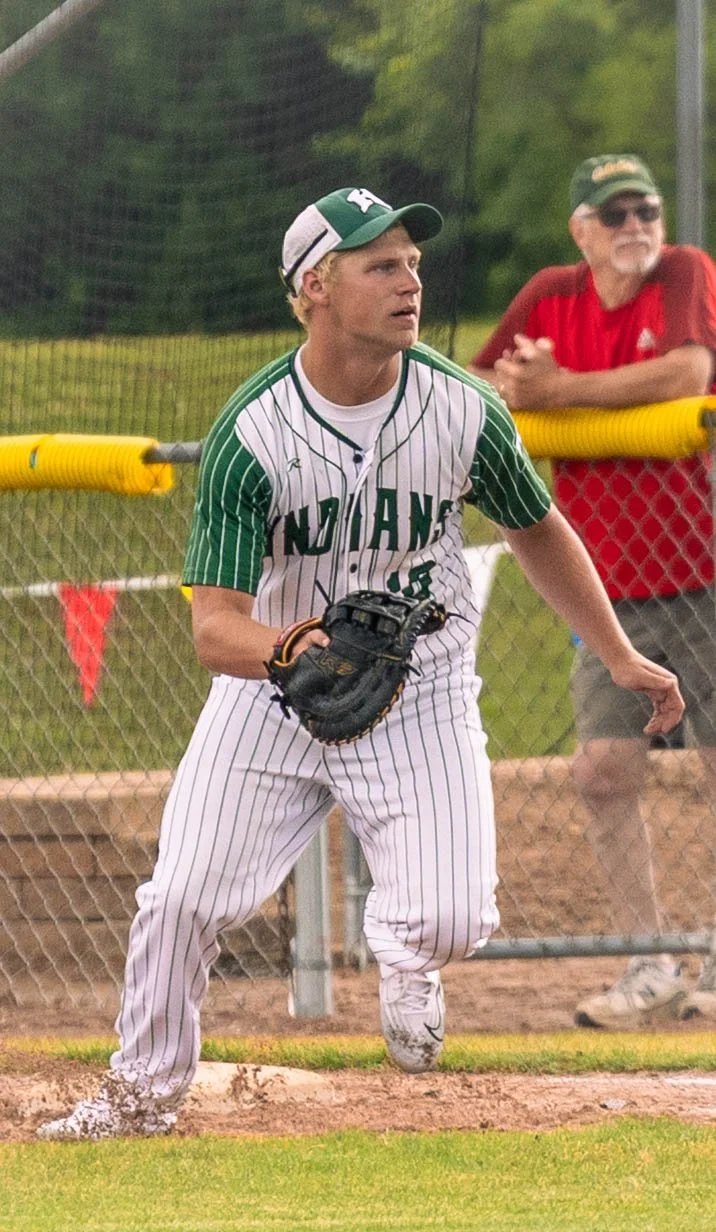 A young male baseball player in a white and green striped uniform, with the word 'Indians' on the front, stands ready on the field with a glove on his left hand, near the dugout fencing. An older man in a red shirt and sunglasses stands behind the fe