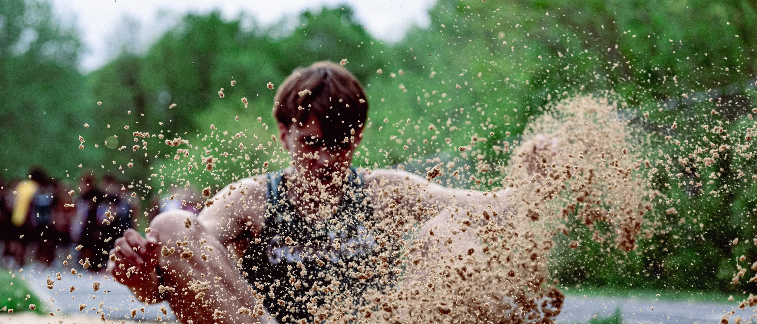A person outdoors throwing a handful of sand or dirt, with a blurred background of trees and other people, in a natural setting on a cloudy day.