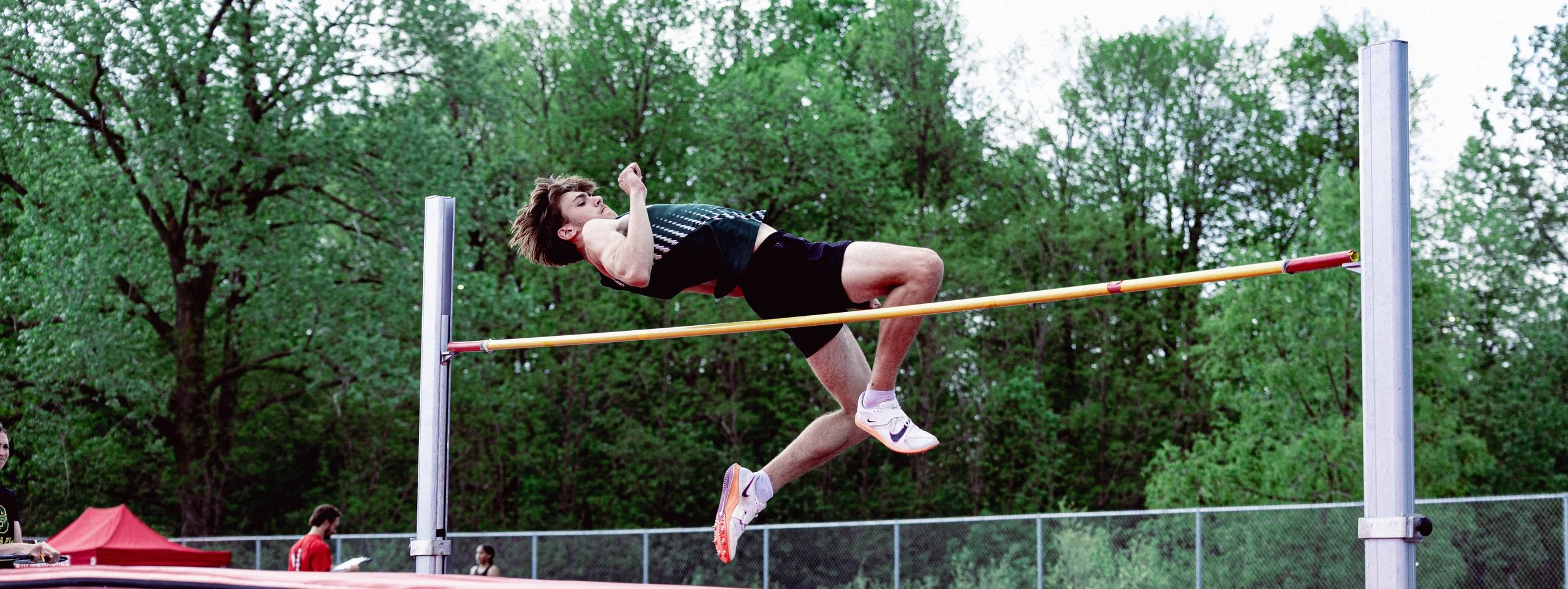 A male athlete competing in high jump, clearing the bar outdoors with trees in the background.