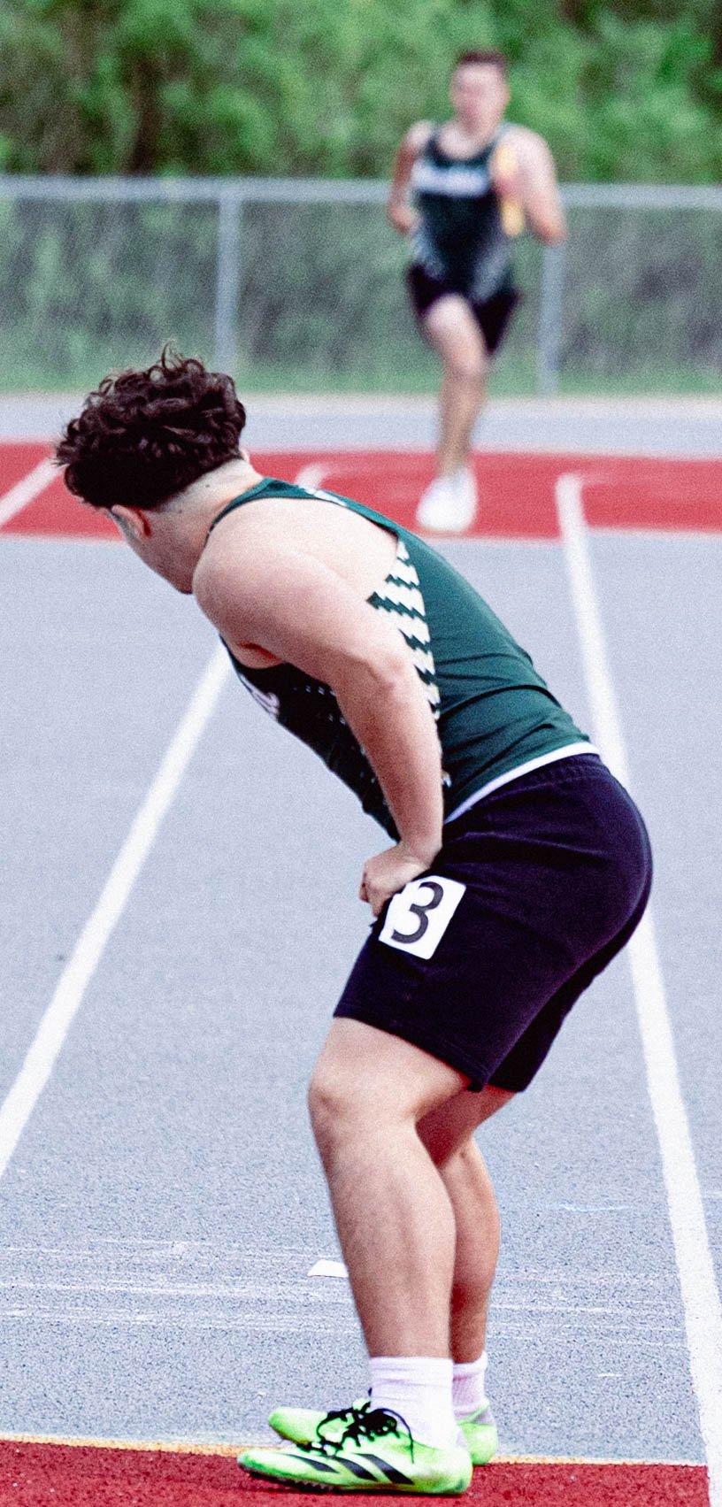 A male sprinter on a track, bending over with hands on knees, wearing a dark green and black athletic outfit and neon green running shoes, with another runner approaching in the background.