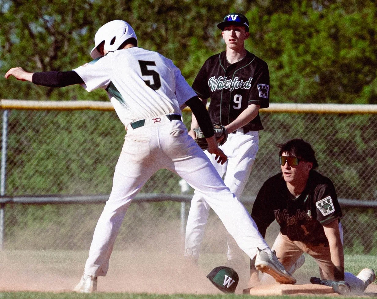 A baseball player in a white uniform with the number 5 reaching for a bag while a player in black with the number 9 and another kneeling player in black observe during a game on a sunny day.