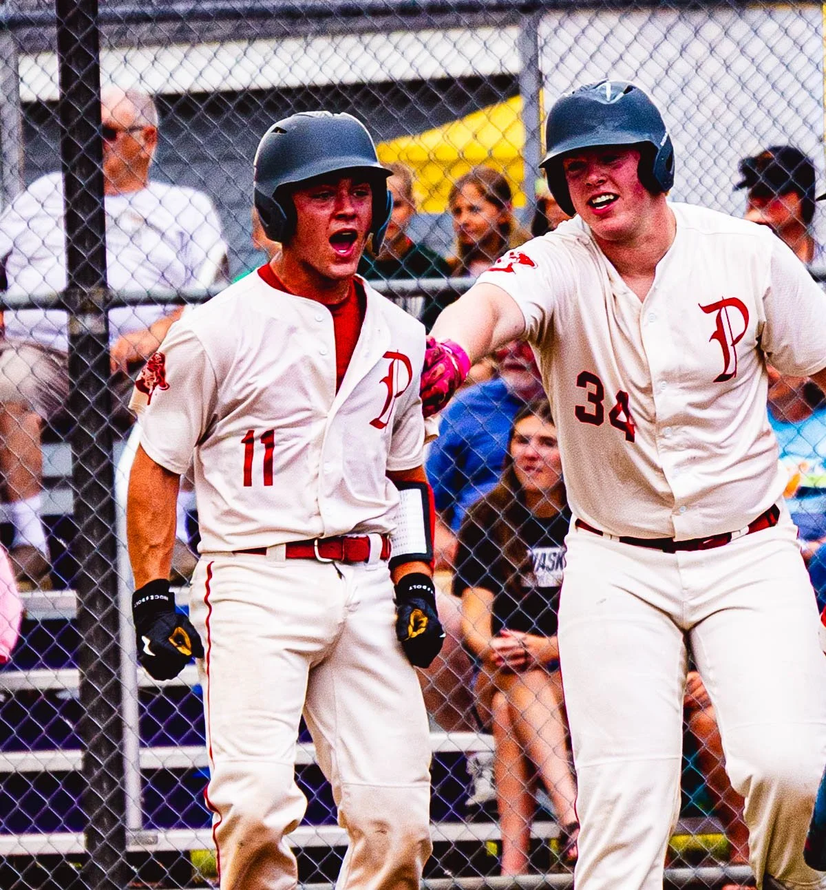 Two baseball players in white uniforms with red accents, wearing helmets, celebrating on the field with a crowd behind a chain-link fence.