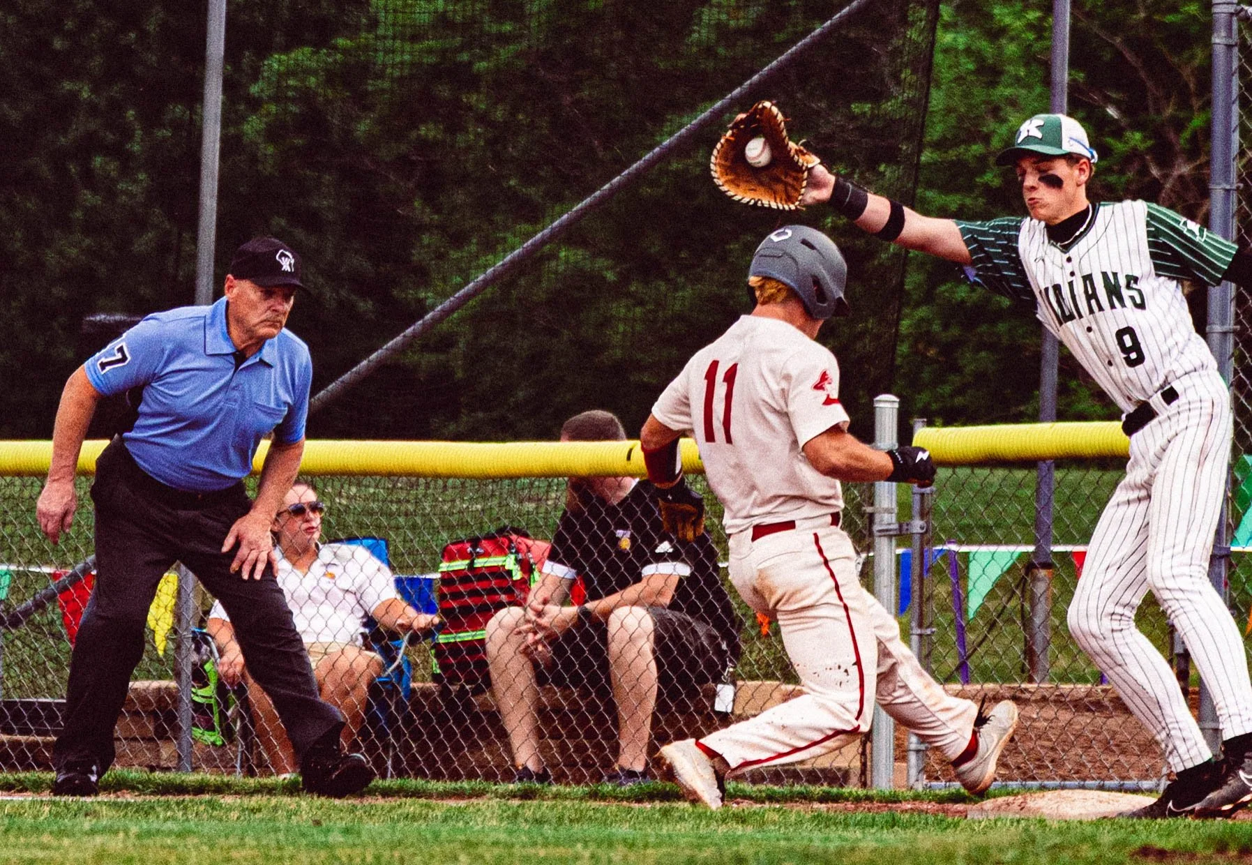 A baseball game with a player running towards the home plate near a catcher and umpire, while spectators watch from the bench behind a chain-link fence. The catcher is wearing gray and red, and the player is in a white uniform with red and black. The umpire is dressed in black and blue. The scene takes place outdoors with trees in the background.