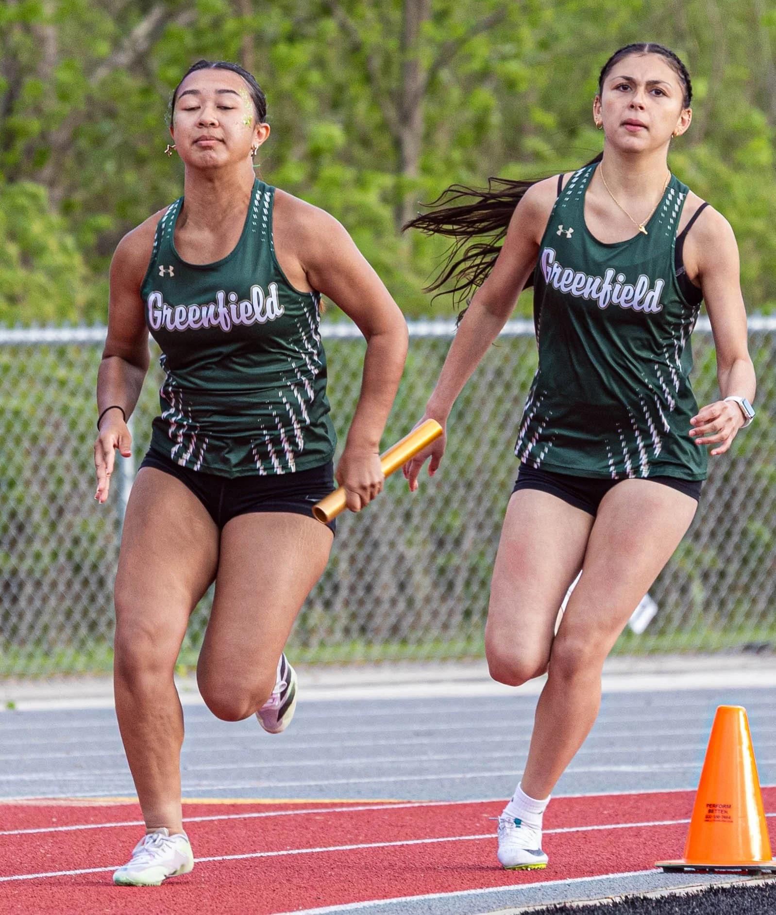Two female athletes running on a track during a race, with orange cones marking the lane, trees in the background, and one athlete holding a relay bat.