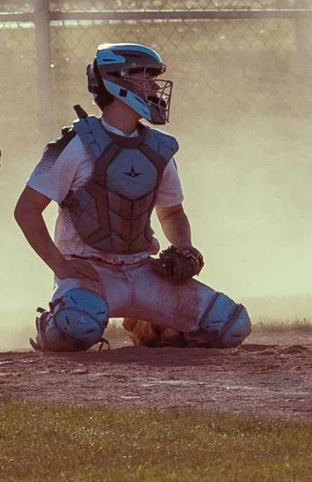 A baseball catcher in gear kneeling behind home plate, wearing a helmet, chest protector, leg guards, and gloves.
